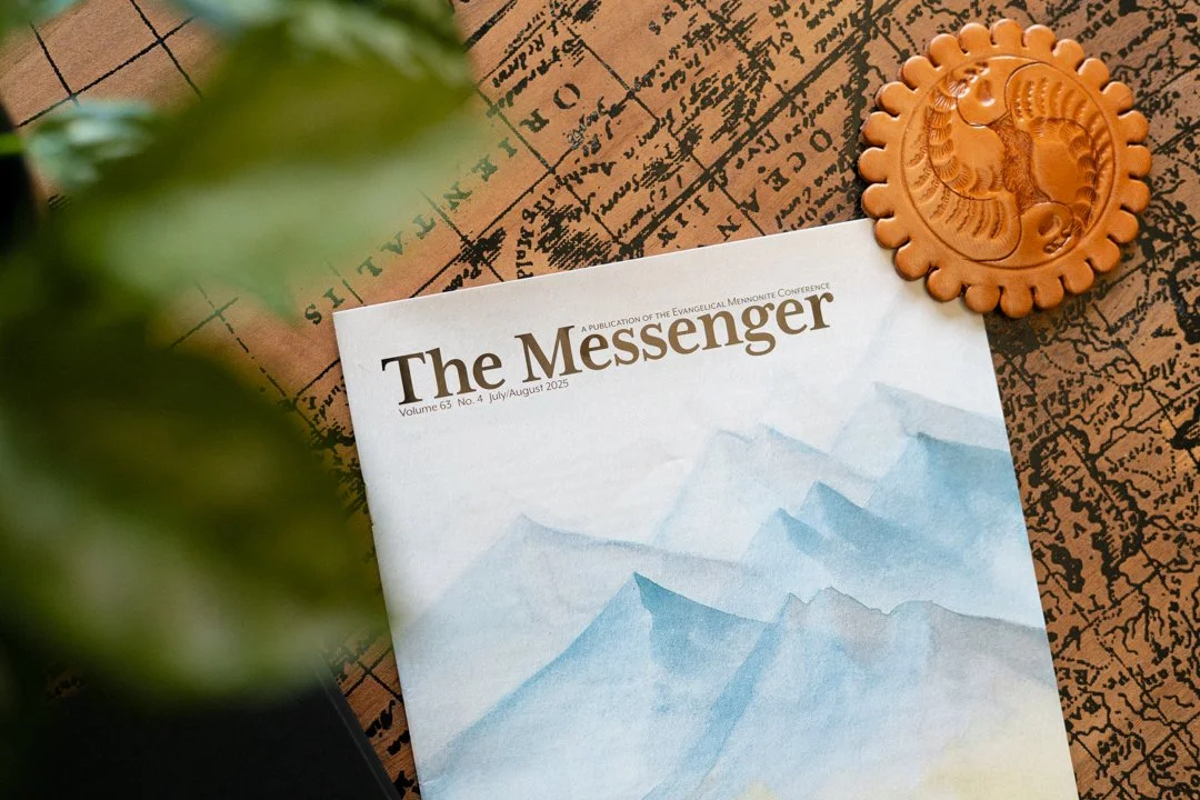 A magazine titled 'The Messenger' with a watercolor painting of mountains on the cover, placed on a vintage map, next to a wooden decorative seal with a carved horse design, partially obscured by a leafy plant in the foreground.