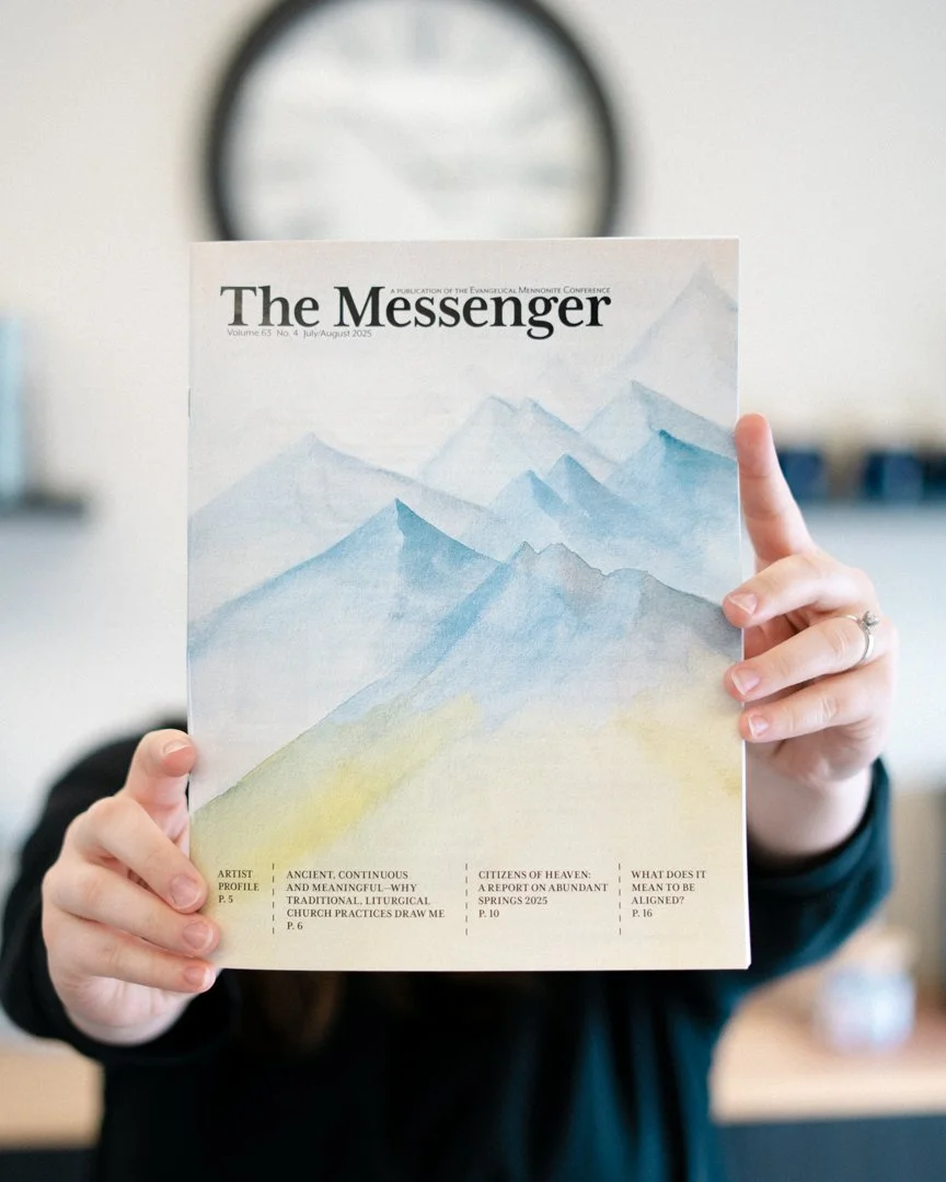 A person holding a magazine titled "The Messenger" with a watercolor painting of blue mountains on the cover.