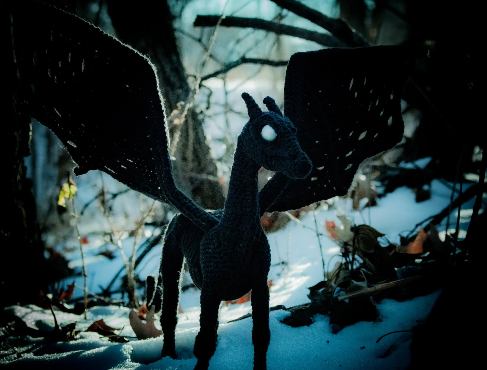 A crocheted Thestrel, a la Harry Potter and the Order of the Phoenix, standing on a shadowed snowbank with its wings on full display.
