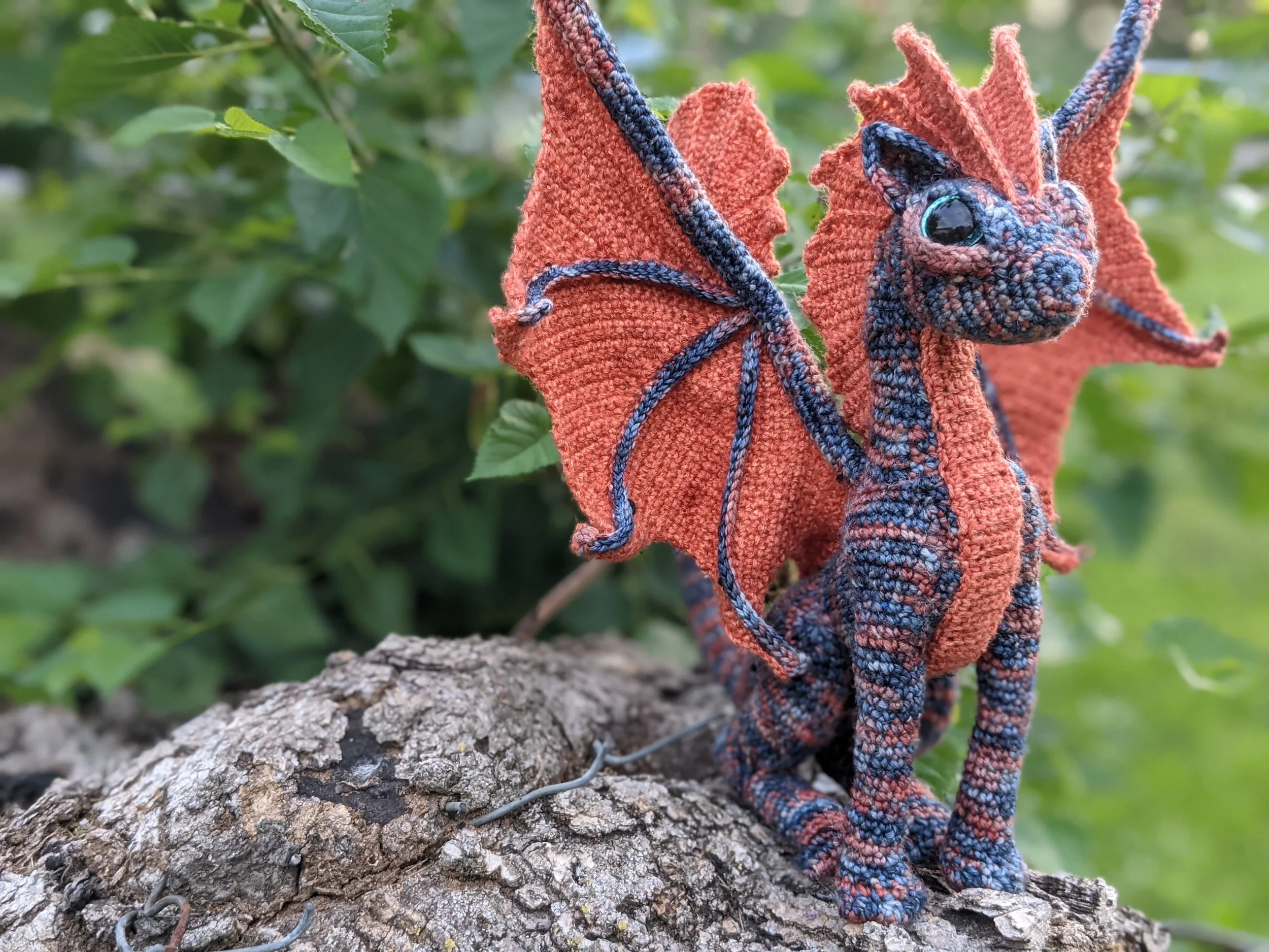 A crocheted cat dragon sitting on a wooden stump among leaves.
