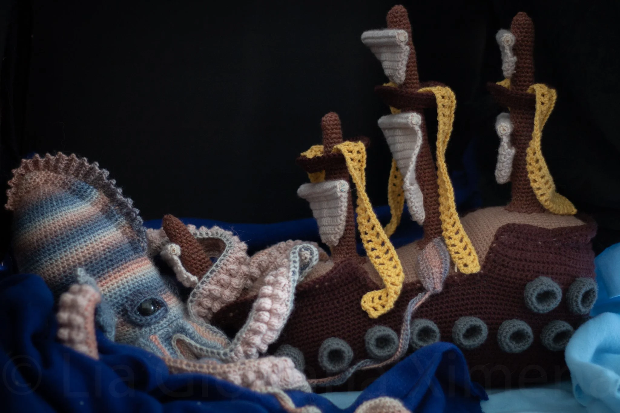 A crocheted kraken and a crocheted pirate ship. The kraken is wrapping its tenacles around the ship's bow.
