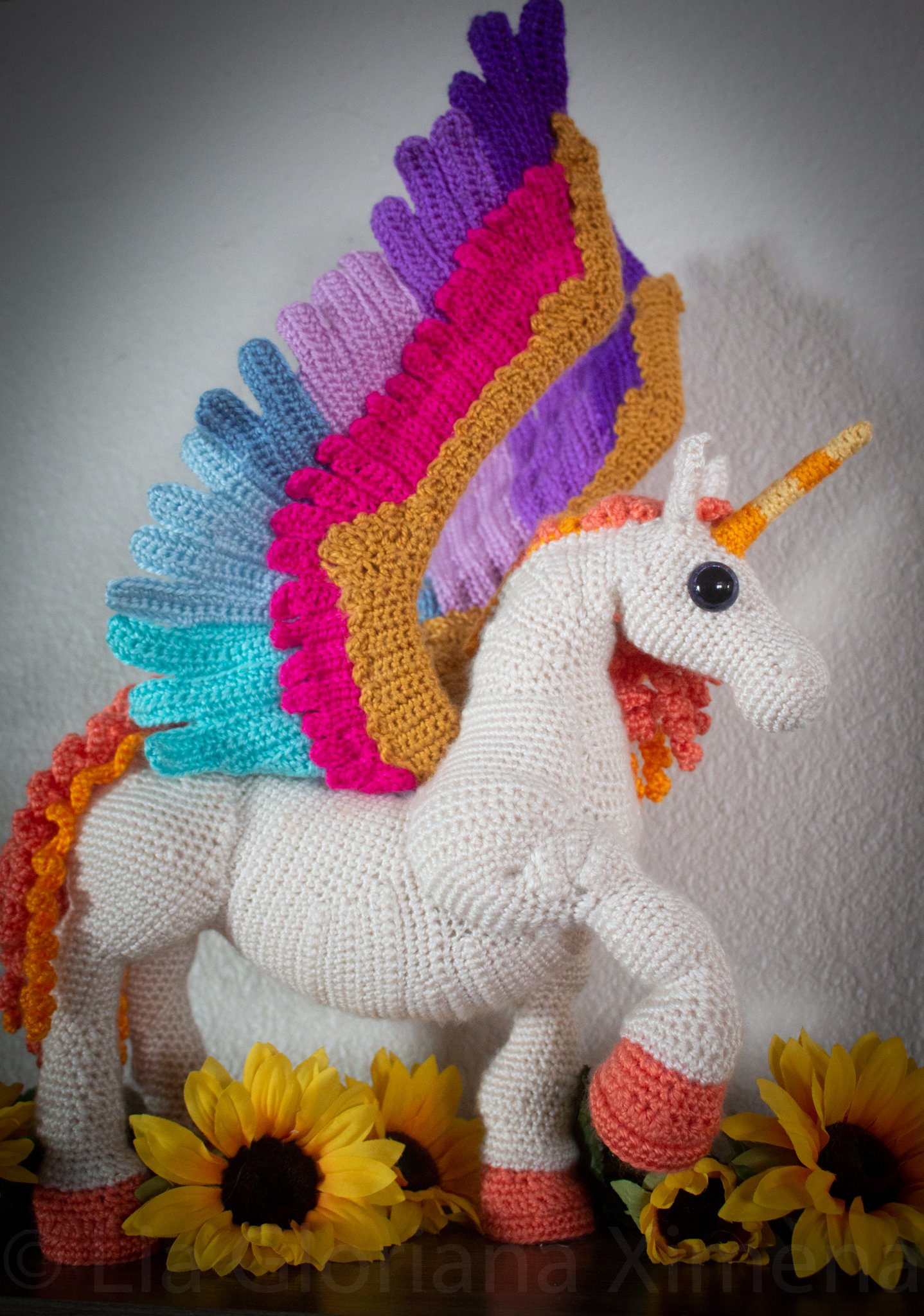 A crocheted multicolored pegasus standing proudly among sunflowers on a shelf.