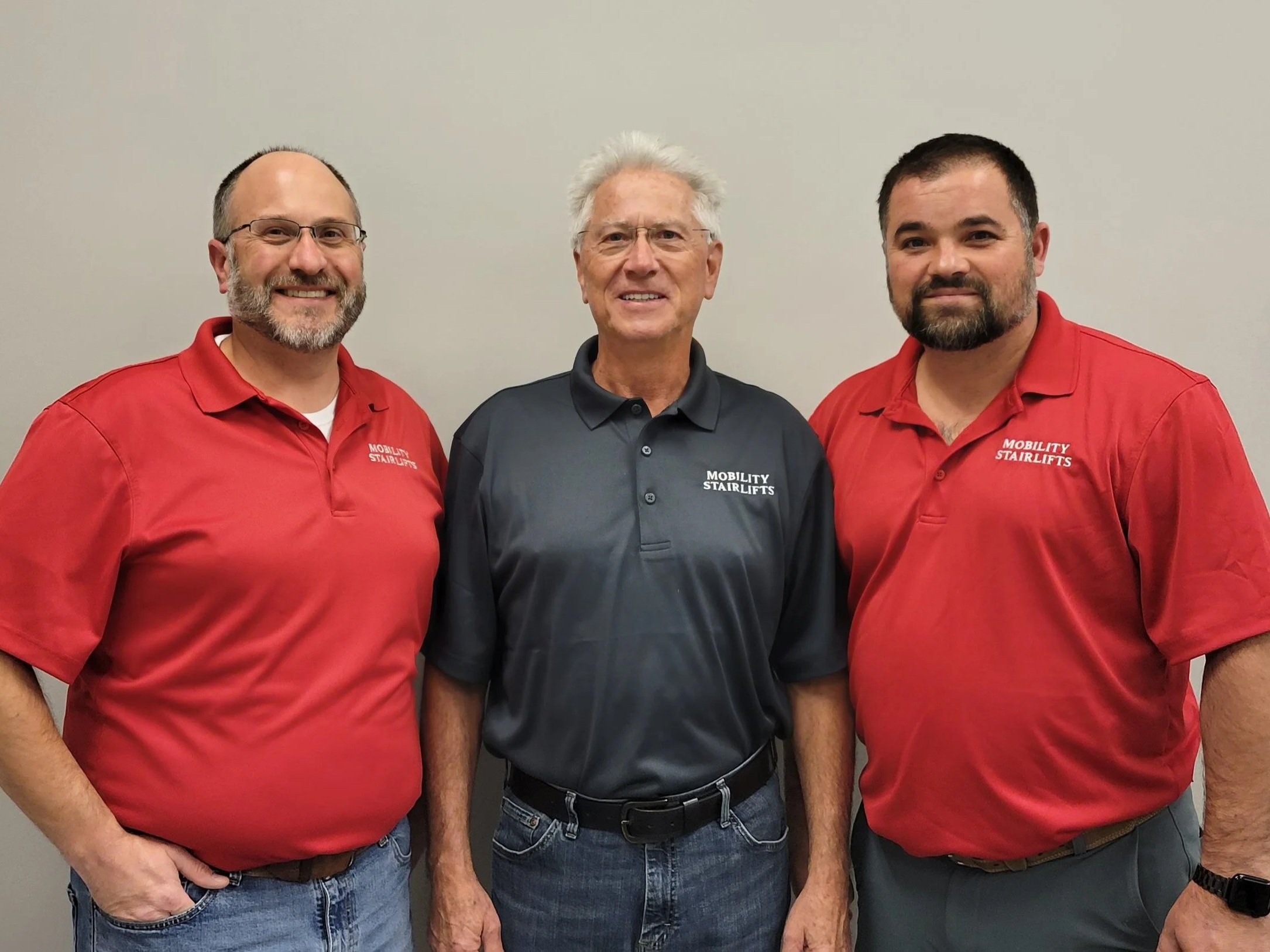 Three men standing side by side against a plain wall, wearing company branded polo shirts with the text 'Mobility Stairlifts'.