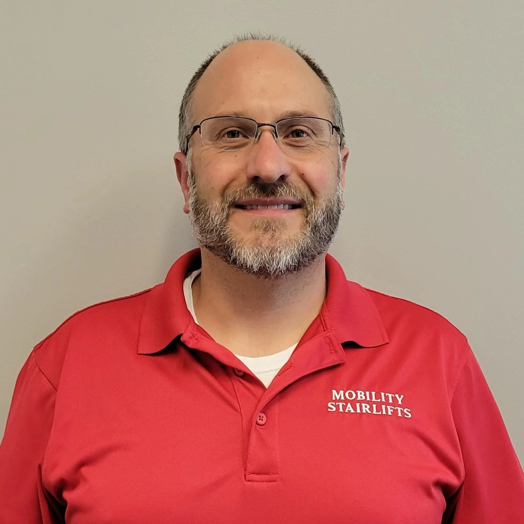 A middle-aged man with glasses and a beard, wearing a red polo shirt with the words 'Mobility Stairlifts' embroidered on it, standing in front of a plain gray wall.