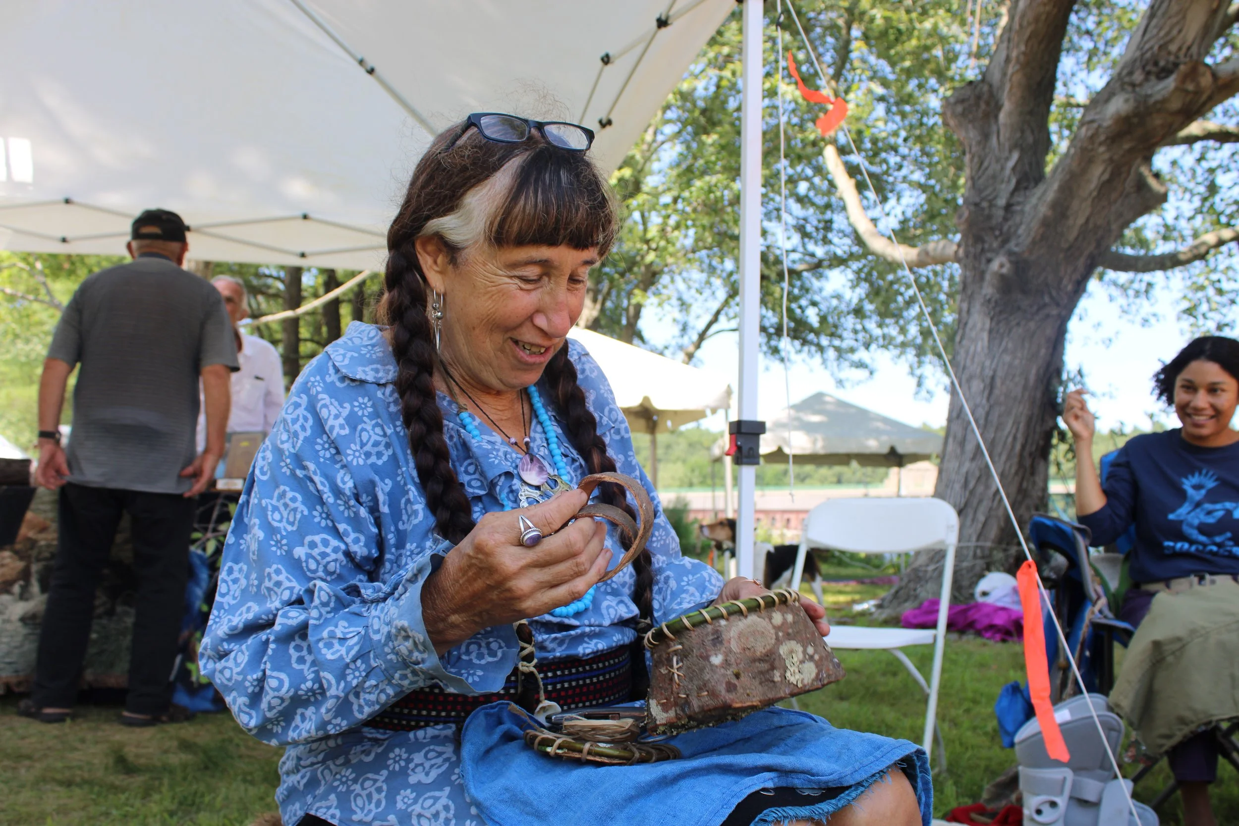 Woman wearing a blue patterned blouse and jewelry, sitting outside at a gathering, holding a bracelet or bangle, with others and trees in the background.