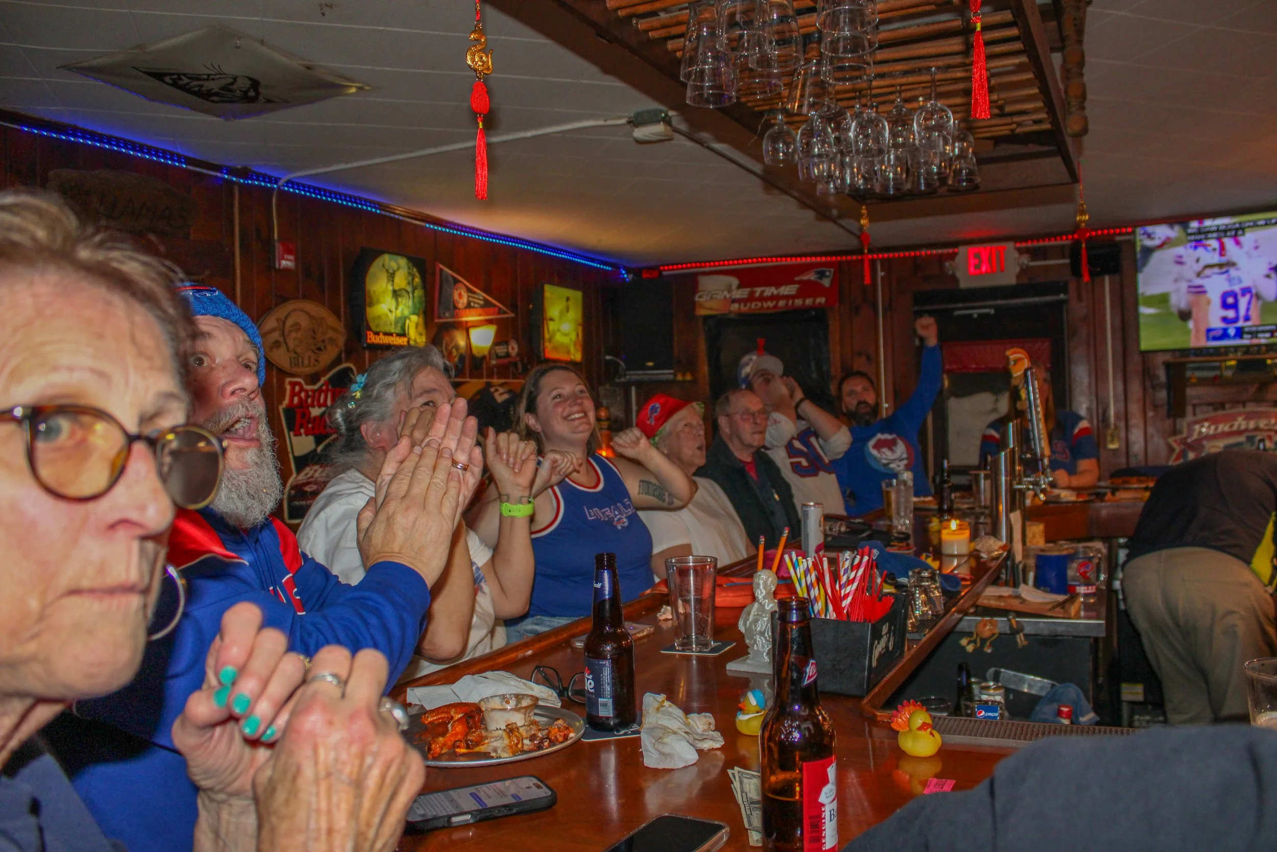 People watching a sports game in a bar with excited expressions, some with hands raised, sitting at a wooden bar counter with drinks and snacks, decorated with sports memorabilia and neon signs.