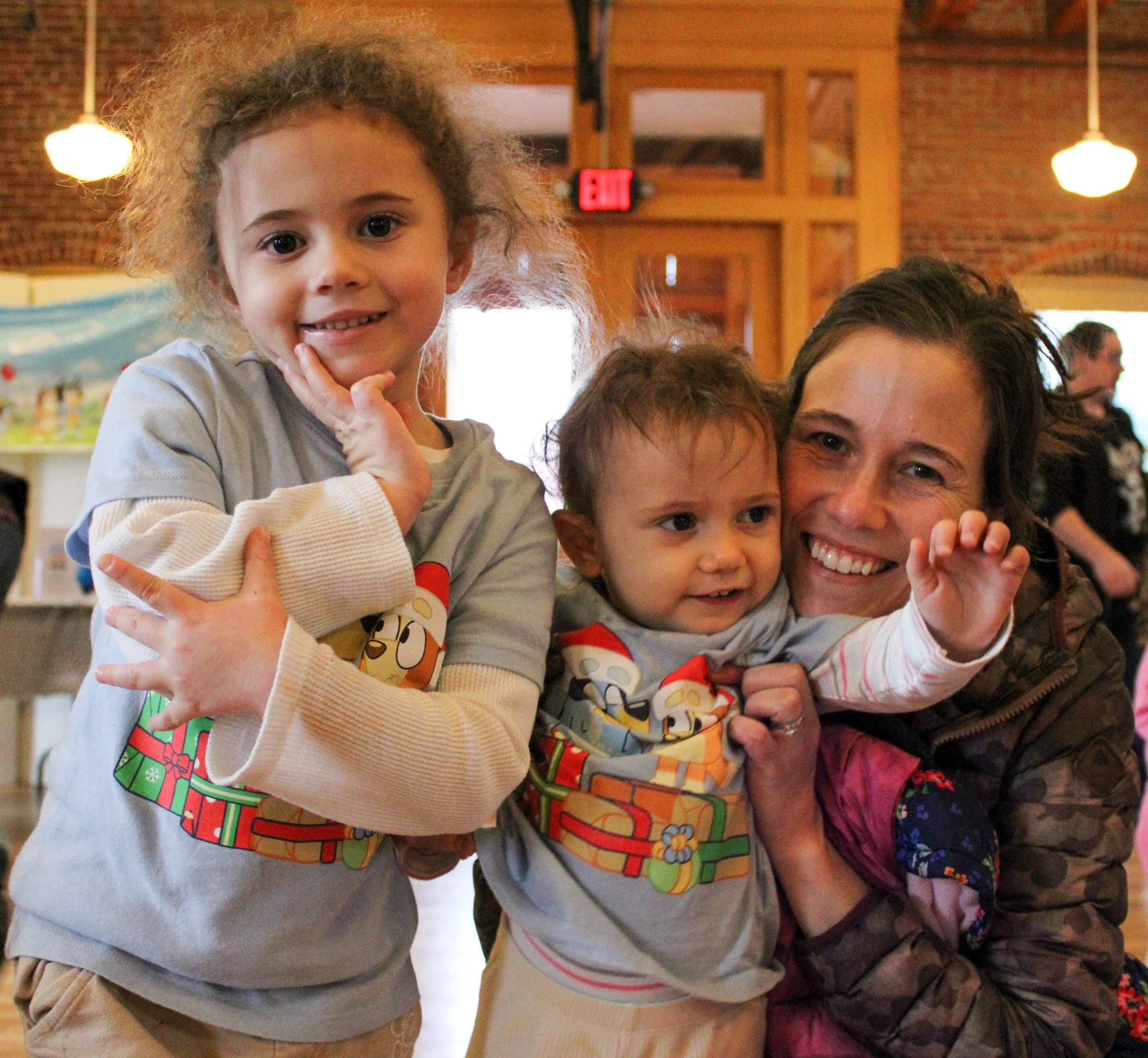 A woman with two young girls in a warmly lit indoor setting, smiling and hugging each other, all wearing festive shirts with cartoon characters.