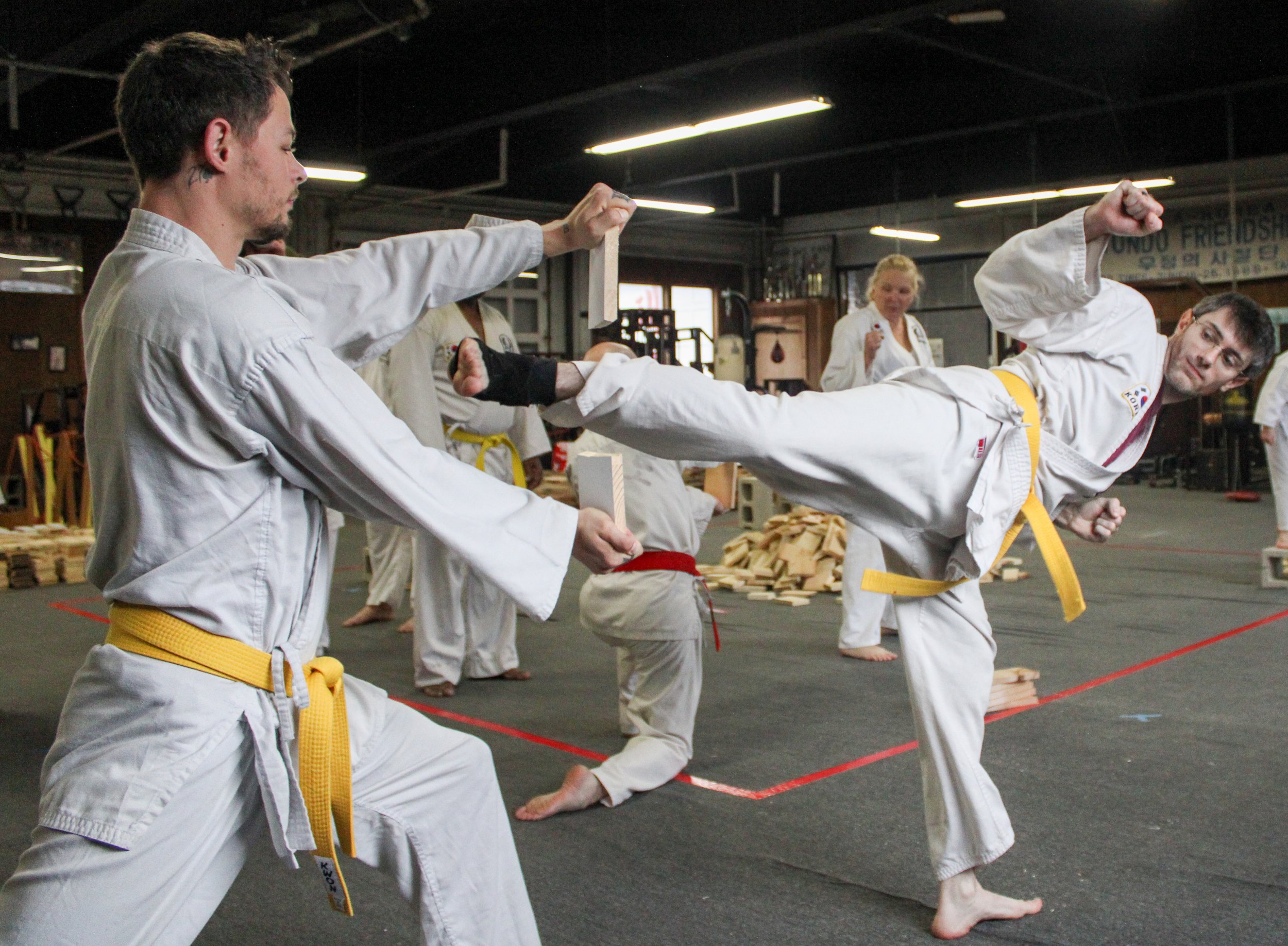 Martial arts class with students practicing Tae Kwon Do. One student is executing a high kick towards a shield held by an instructor, while other students observe in the background.