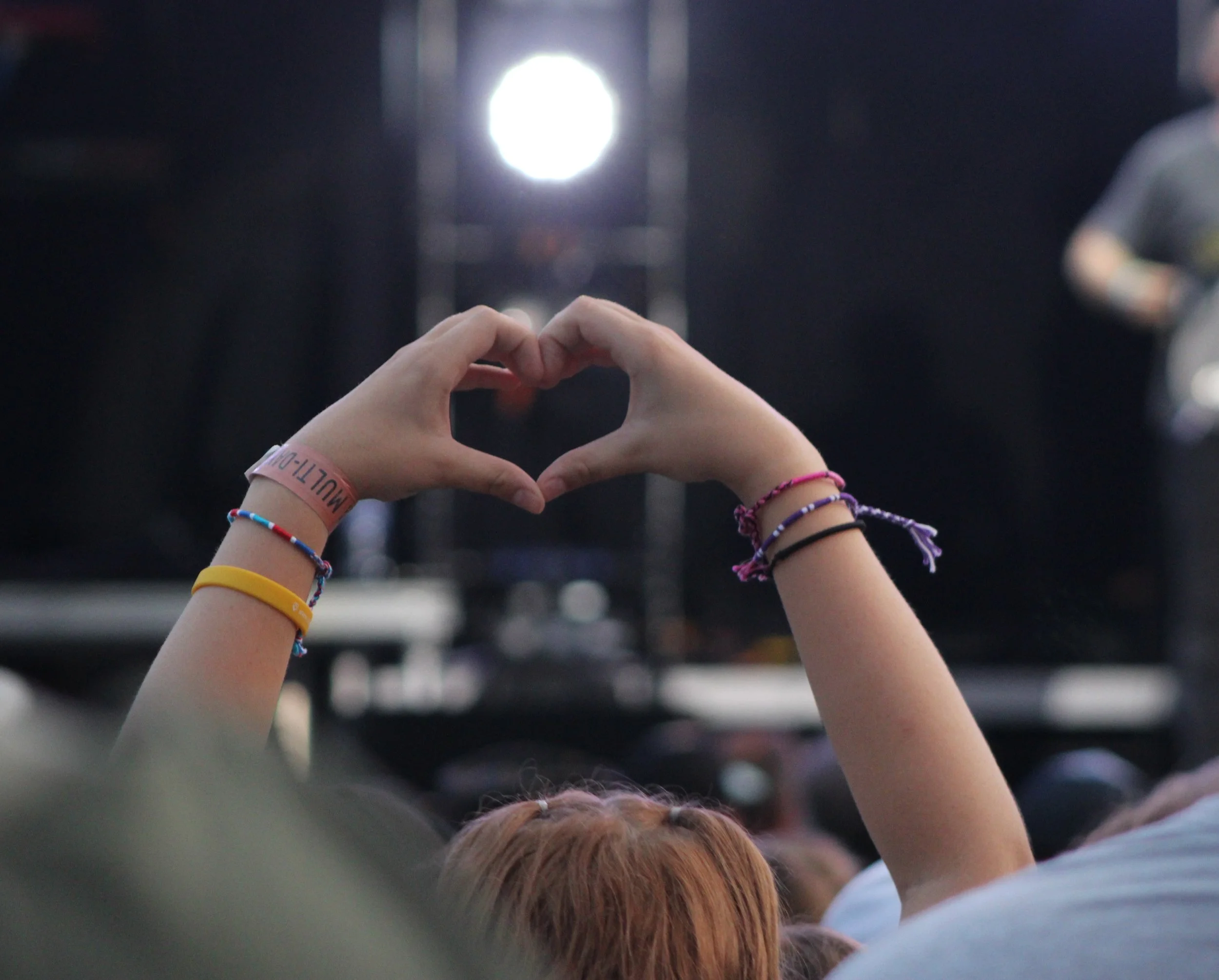 A person at a concert creating a heart shape with their hands, with stage lights in the background.