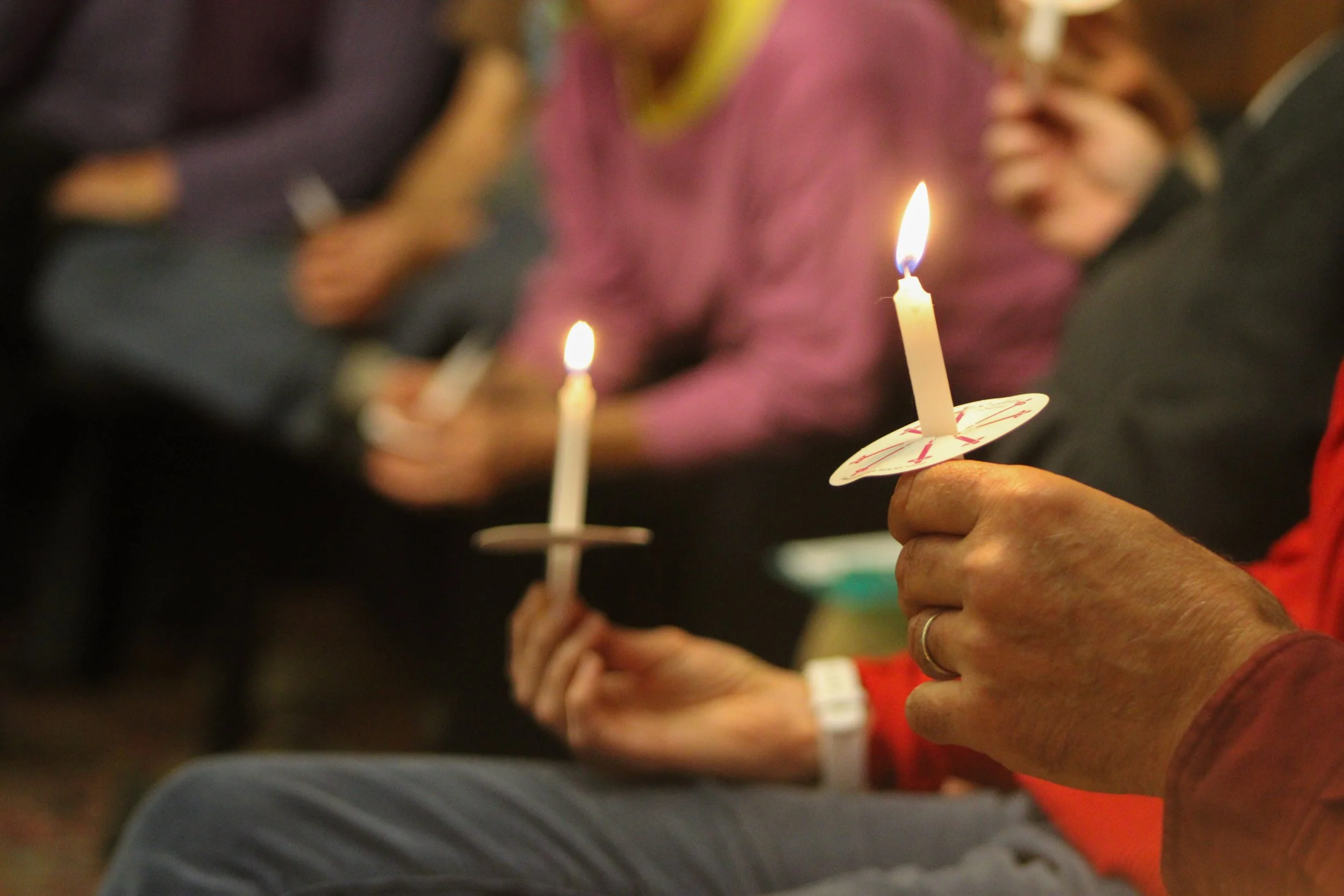 People holding lit candles with paper holders during a gathering.