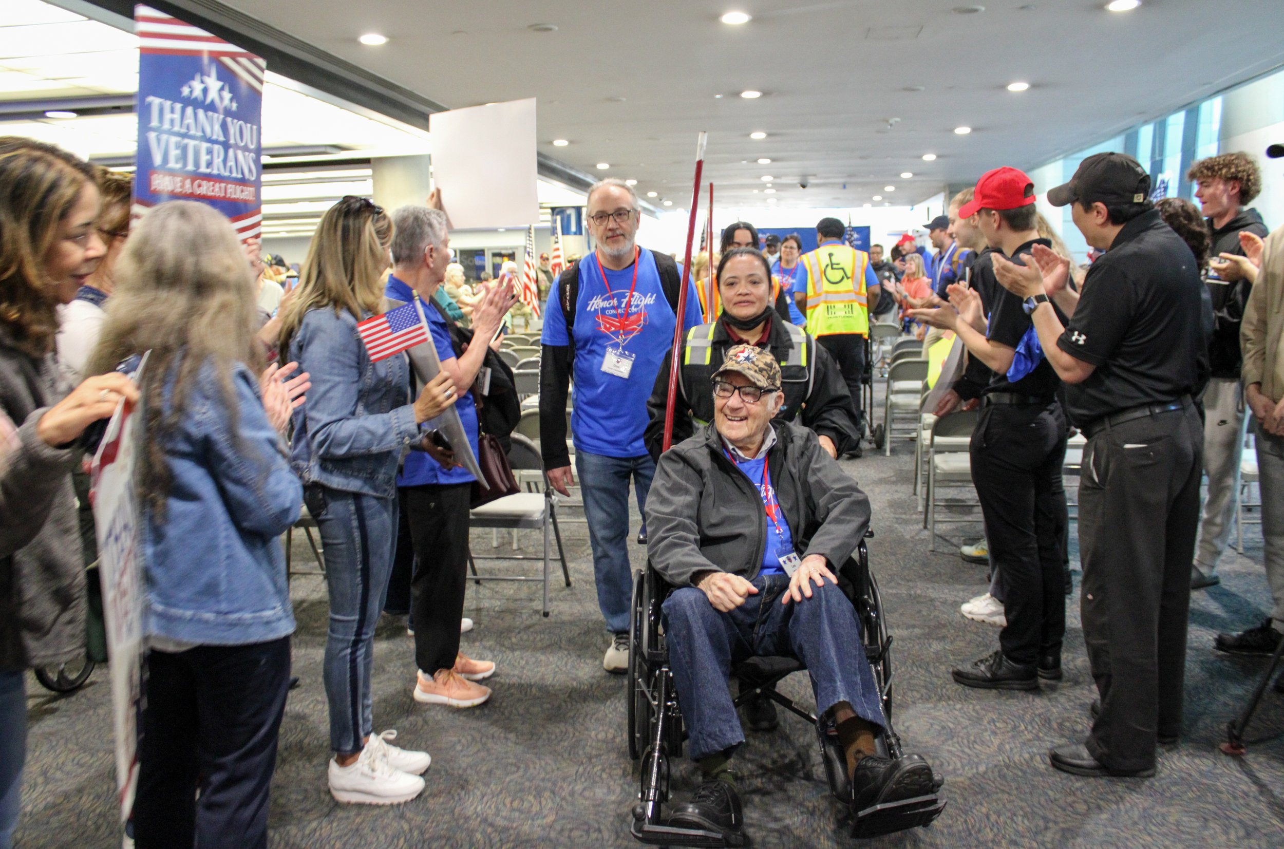 A crowd of people at an airport greeting a veteran in a wheelchair, holding American flags and clapping. The veteran is smiling, wearing glasses, a camouflage cap, and a gray jacket. The scene appears to be part of a welcome or appreciation event.