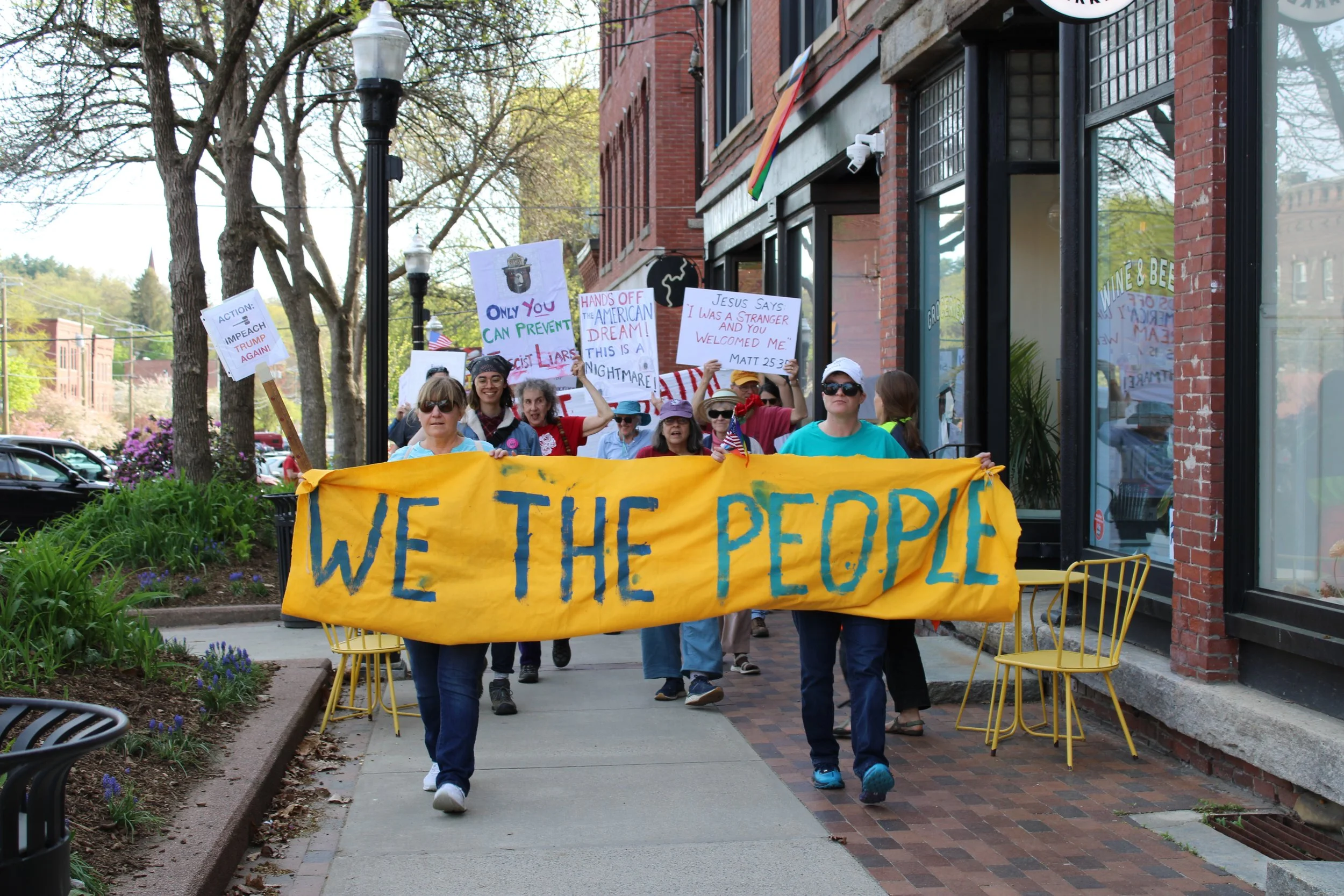 Group of people participating in a march or protest on a sidewalk, holding a large yellow banner that says "WE THE PEOPLE" in blue letters. Some individuals are holding signs with various messages, and there are trees and shops in the background.