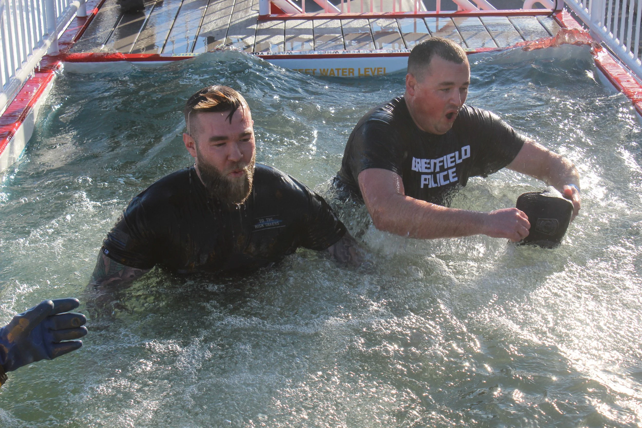Two men participate in a water rescue training exercise in a pool. One wears a black shirt labeled 'Brestfield Police' and the other a shirt with a patch. They are submerged up to their chests in the water with a metal railing and a sign that reads '