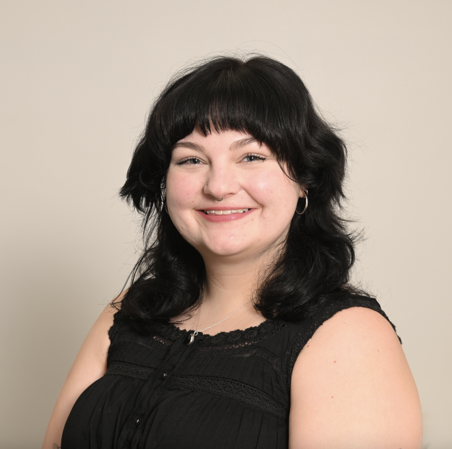 A woman with black hair, wearing a sleeveless black top and jewelry, smiling at the camera against a plain background.