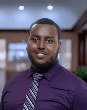 A man with short hair and a beard wearing a purple dress shirt and striped tie smiling indoors.