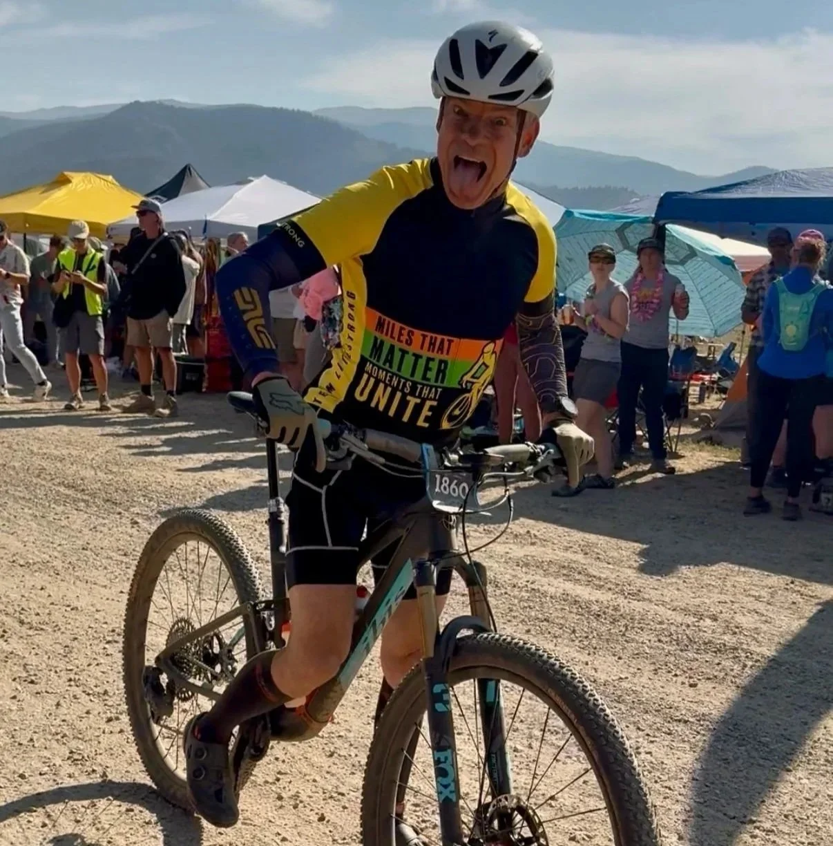 Person on a mountain bike in yellow and black bike jersey with aid tents in the background