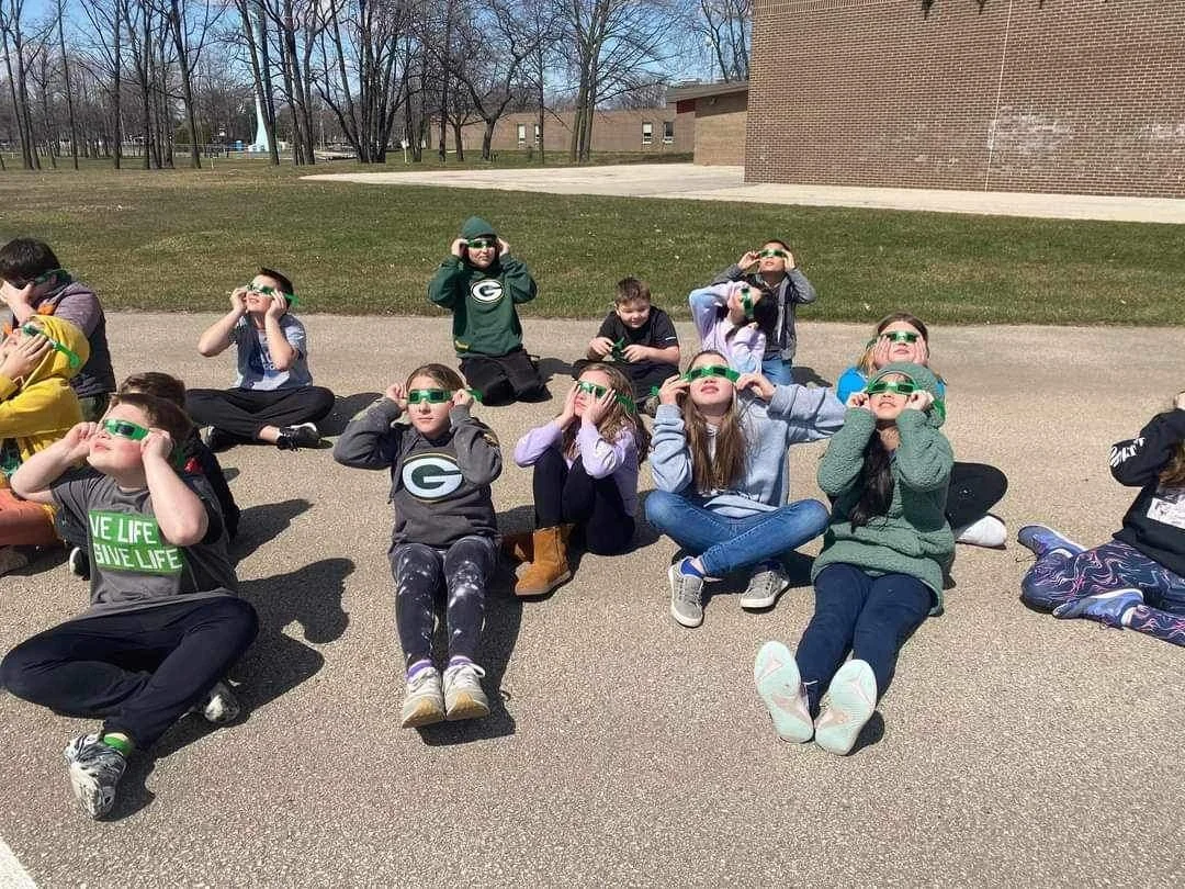 A group of children sitting on the ground outside, wearing solar eclipse glasses, some covering their ears or eyes, on a sunny day with a grassy field and trees in the background.