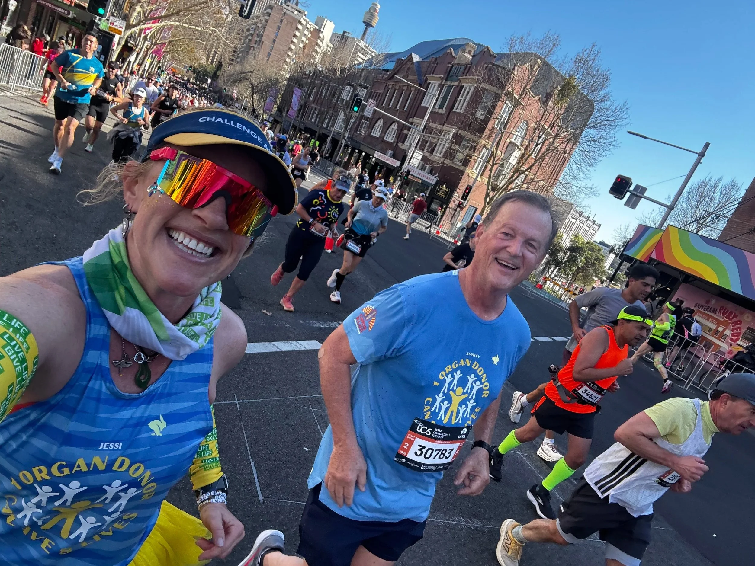 Group of marathon runners participating in a race on a city street, smiling and wearing athletic gear, with buildings and a crowd in the background.