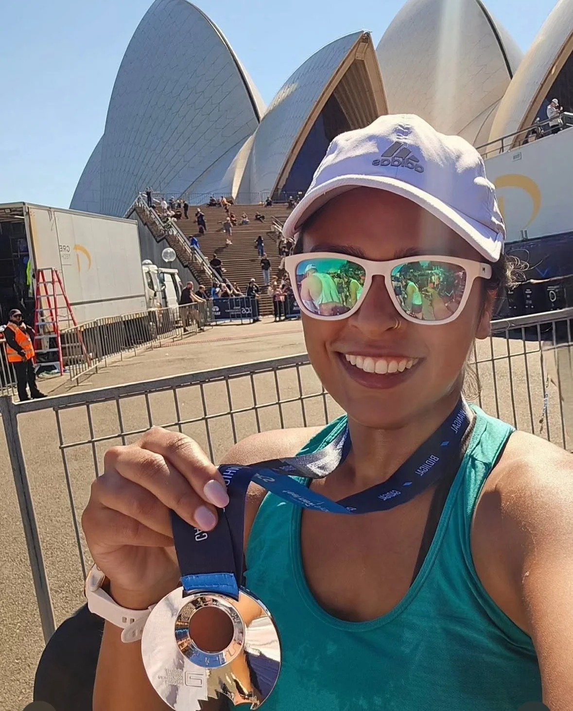 A woman smiling outdoors in front of the Sydney Opera House, holding a silver medal. She is wearing sunglasses, a white cap, a turquoise tank top, and a smartwatch. The background shows steps leading up to the opera house and several people.