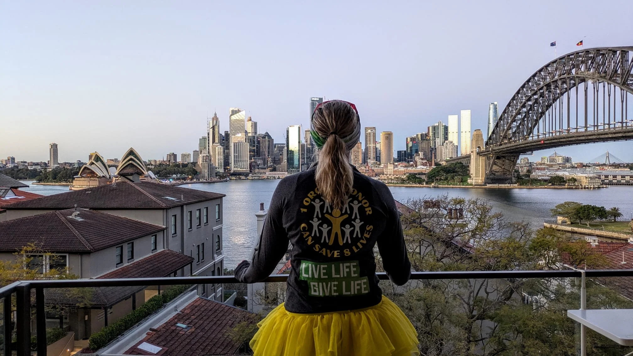 A woman with a yellow tutu and black hoodie stands looking over Sydney Harbour, featuring the Sydney Opera House and Harbour Bridge, in Australia.