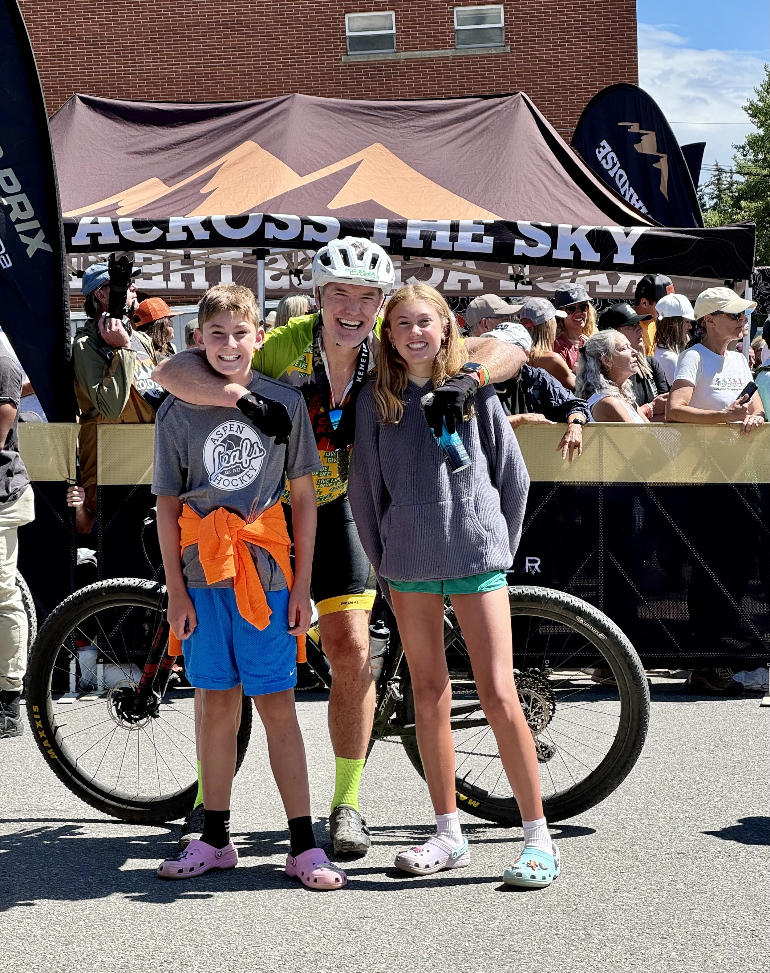 A smiling man and girl on a mountain bike at an outdoor event, the girl is holding a medal, with others in the background near parked vehicles.