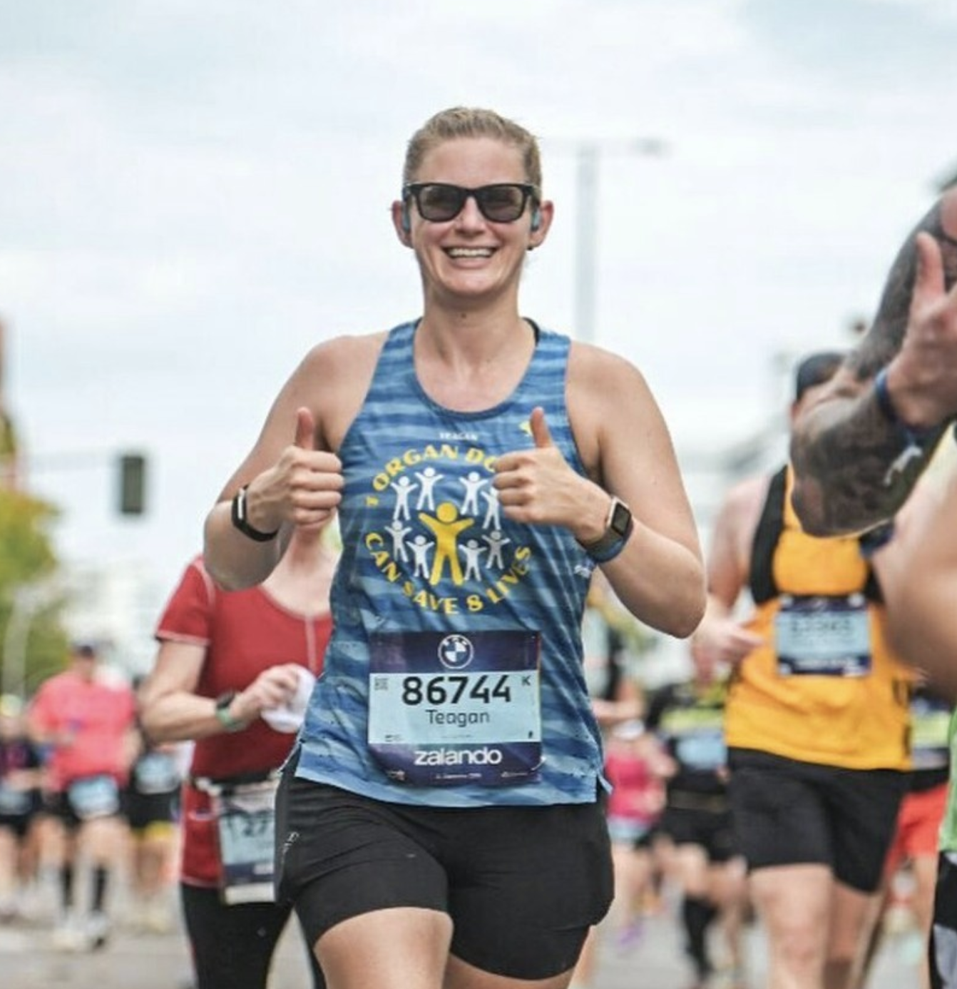 Woman running in a marathon, smiling, wearing sunglasses, a blue tank top with a community message, black shorts, and a race bib numbered 86744.