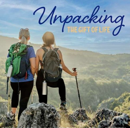 Two women with backpacks hiking on a trail overlooking a scenic valley with rolling hills and a partly cloudy sky. The text says 'Unpacking the Gift of Life'.