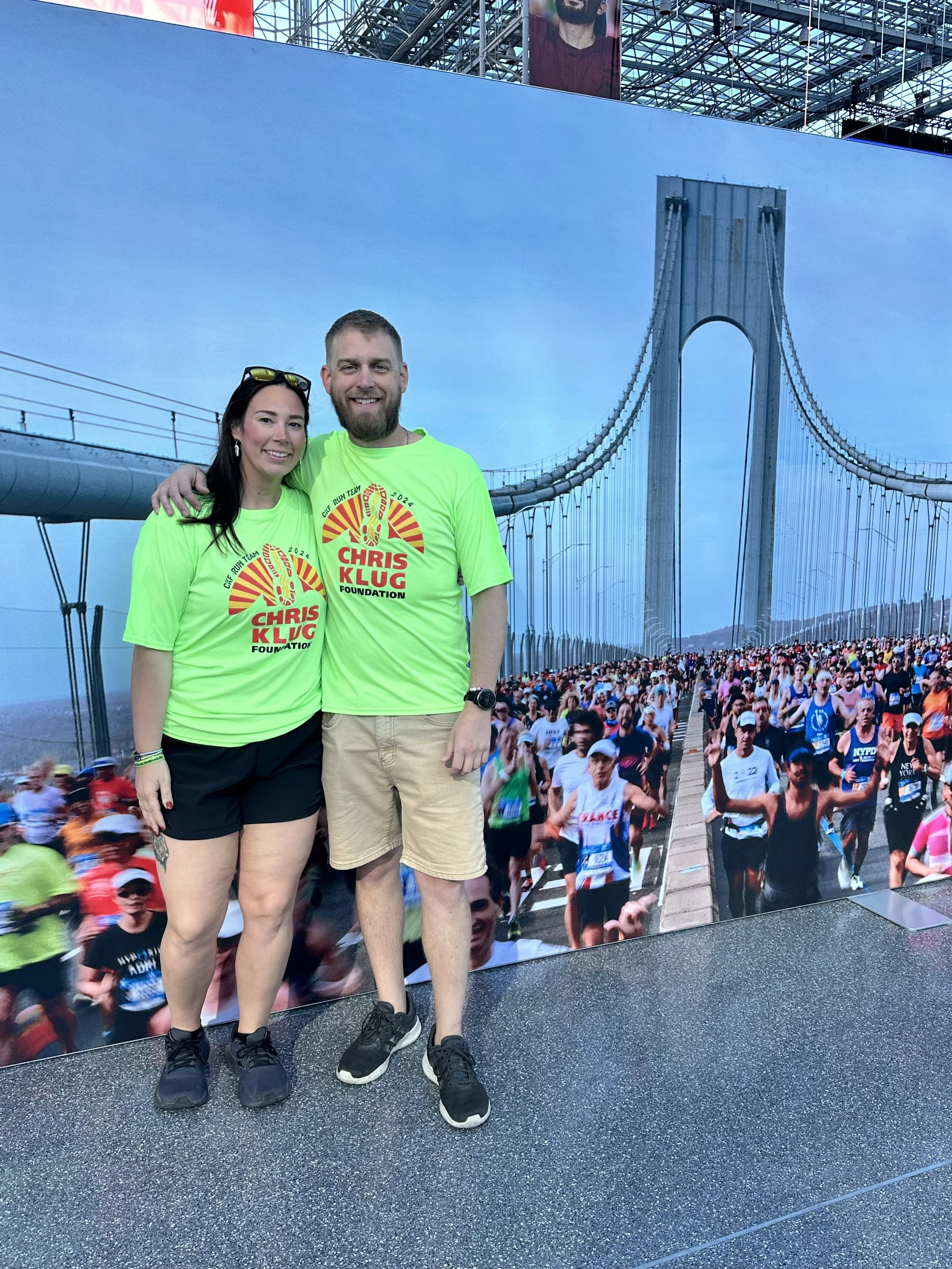 Two people in bright green shirts with 'Chris Klug Foundation' logo standing in front of a large wall displaying a marathon scene on a bridge, with a crowd of runners.