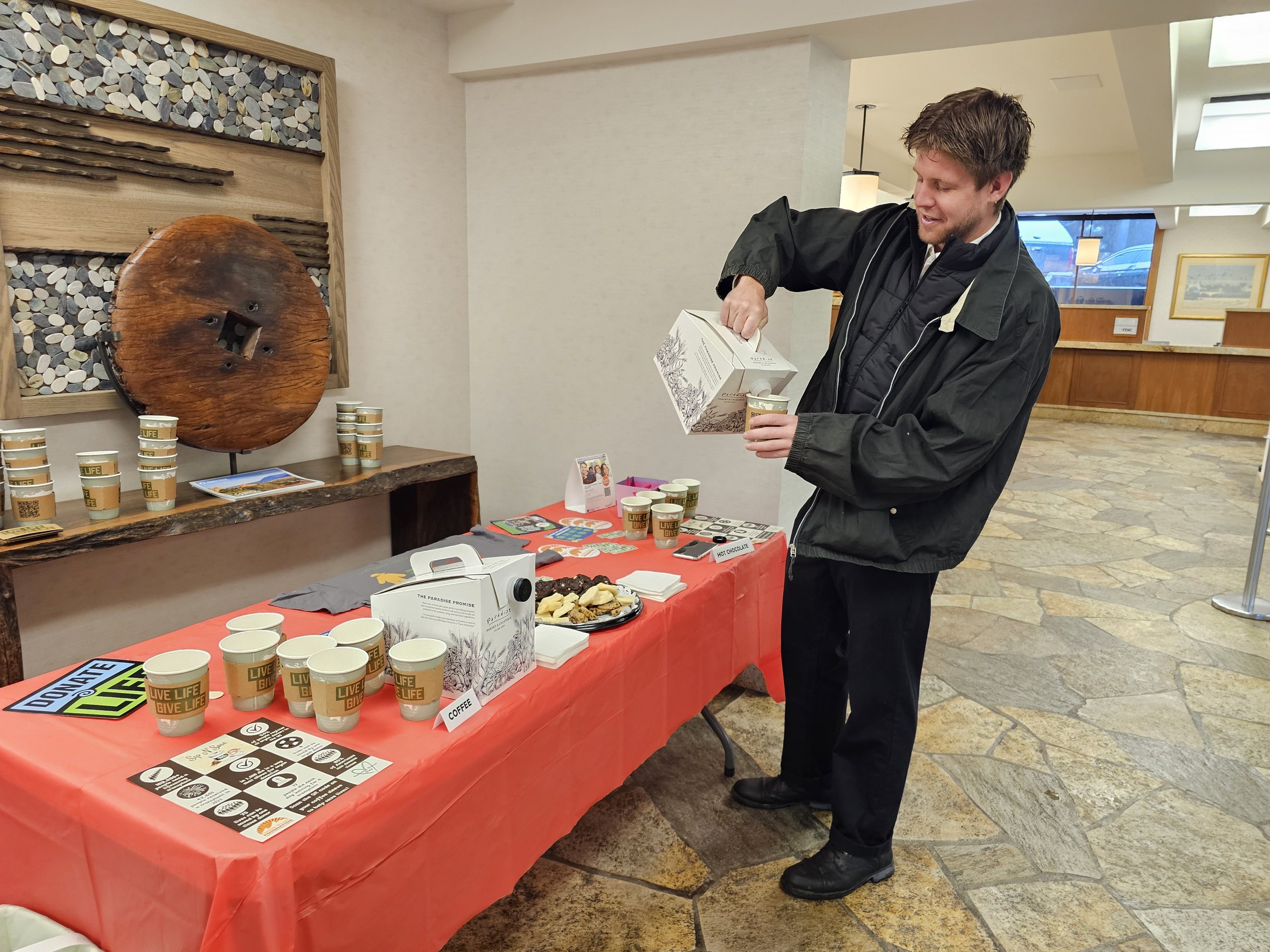 A man in black jacket pouring hot chocolate from a carton into a cup at a cafe table with coffee cups, snacks, and promotional materials, in a cozy interior space.