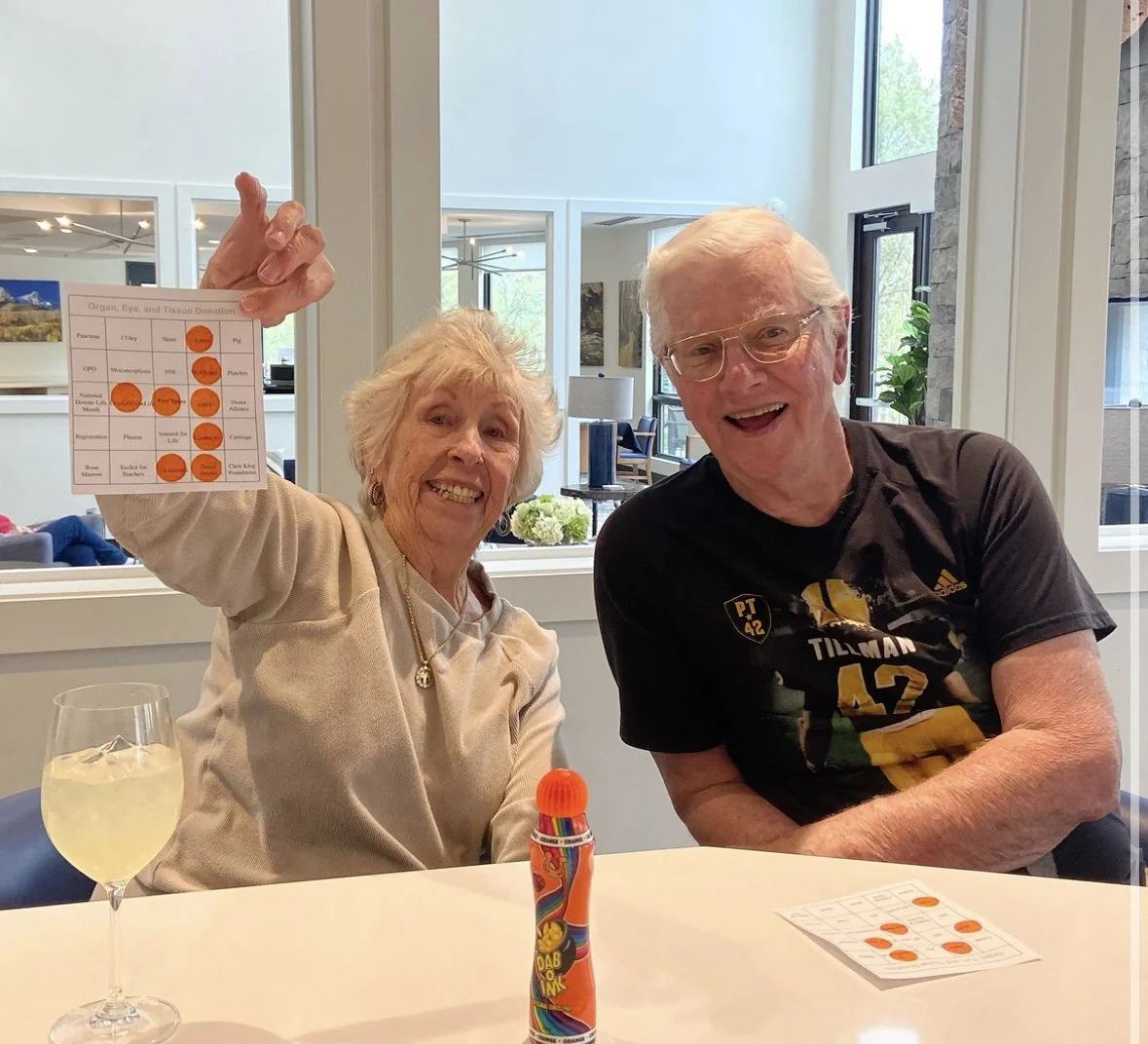 Two elderly people sitting at a table, smiling. The woman is holding up a card with orange circles, and the man is wearing a Black and yellow sports jersey. There is a glass with a light-colored drink and a colorful bottle on the table, with windows and a bright interior in the background.