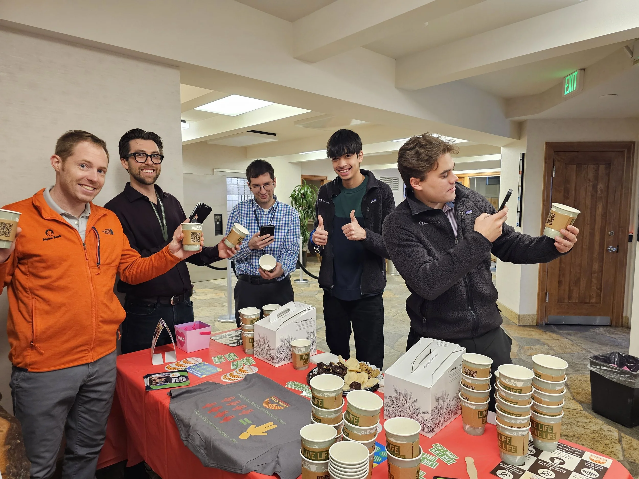 Group of six young men at a table with coffee cups, snacks, and promotional items, in an indoor setting.
