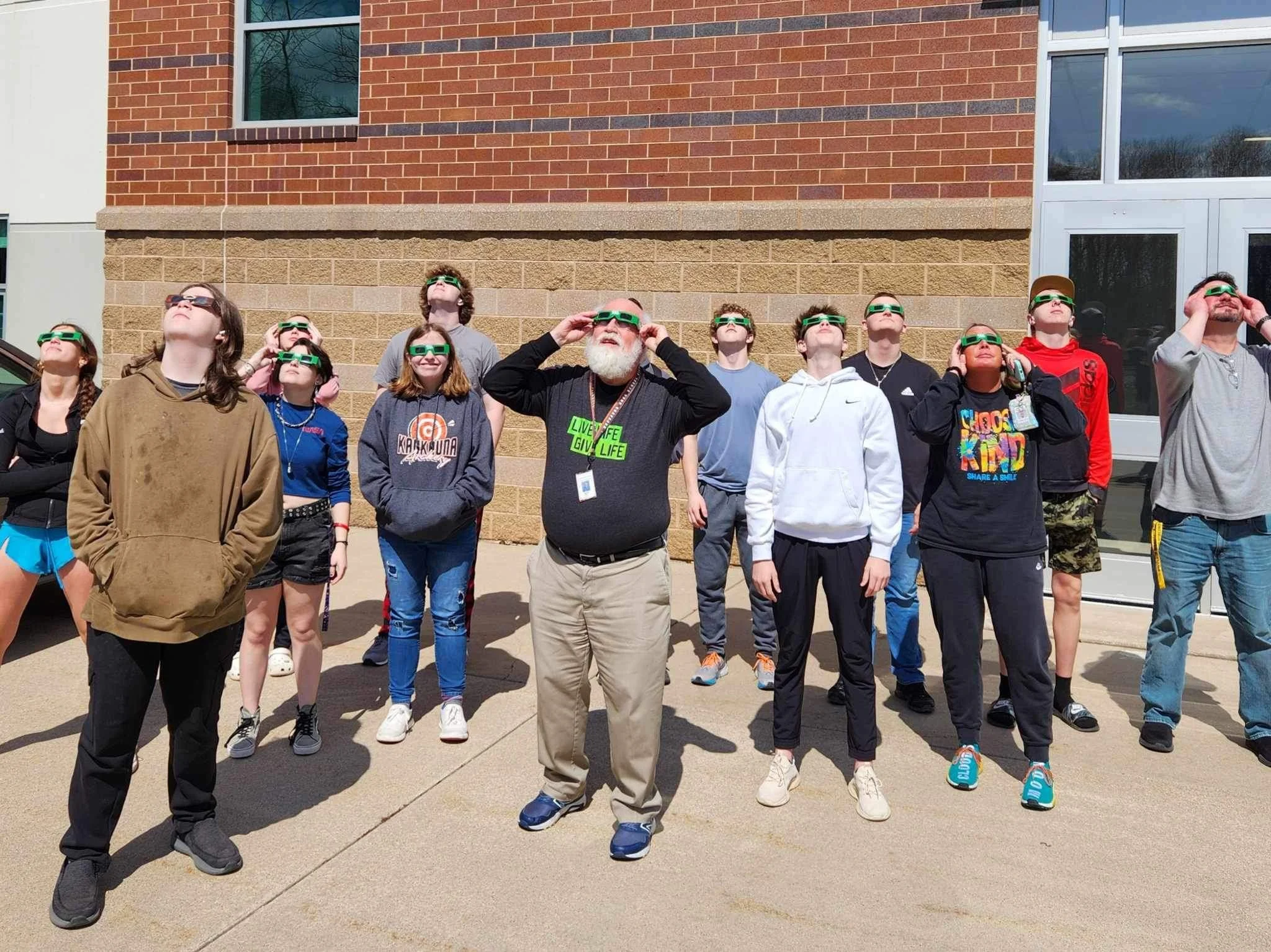 Group of people standing outside, looking up at the sky while wearing green solar eclipse glasses.