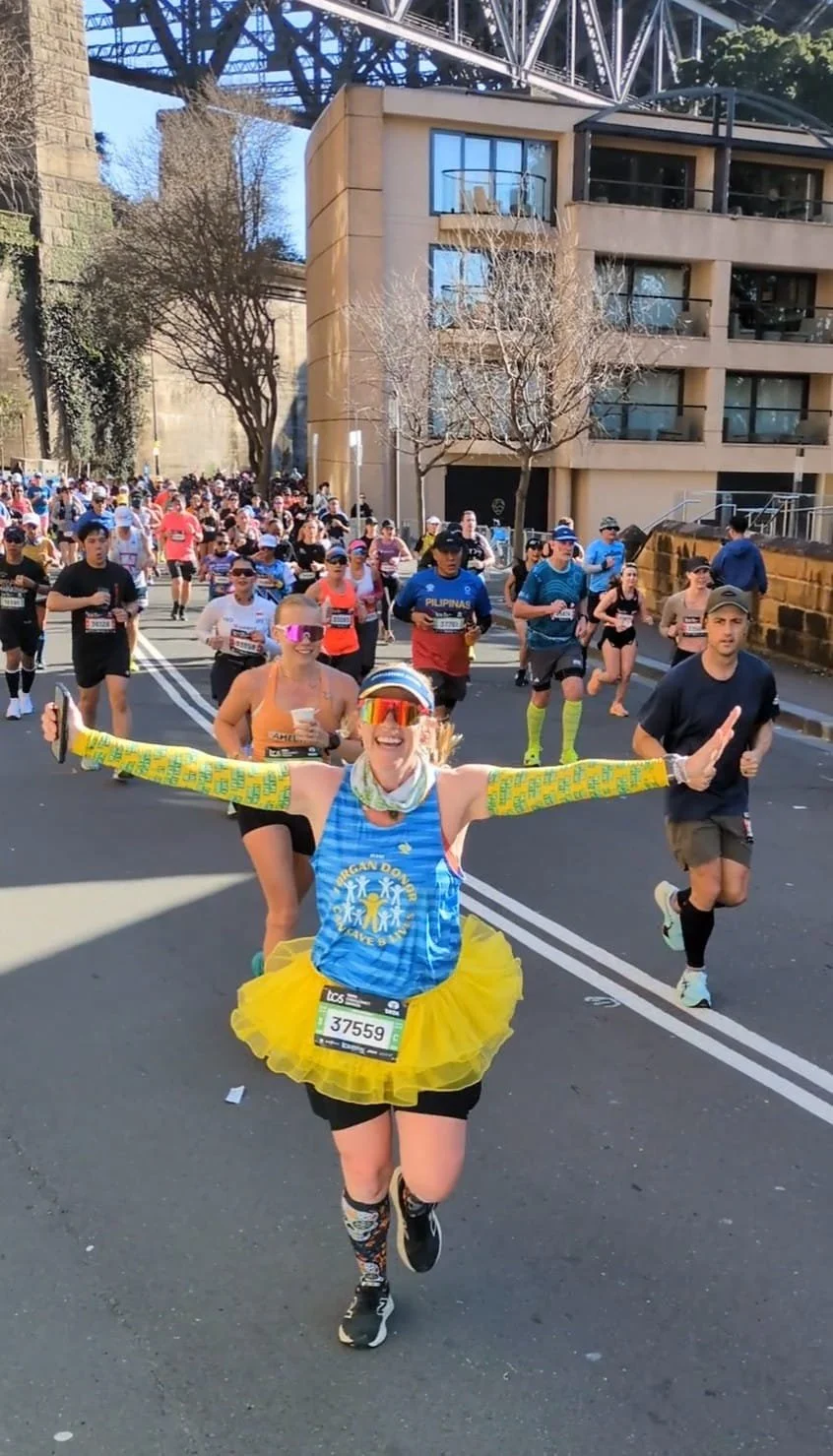 A female runner in colorful attire, including a yellow tutu, long yellow arm sleeves, and a blue tank top, smiles and waves as she crosses the finish line of a marathon. She is surrounded by other participants and spectators on a city street.