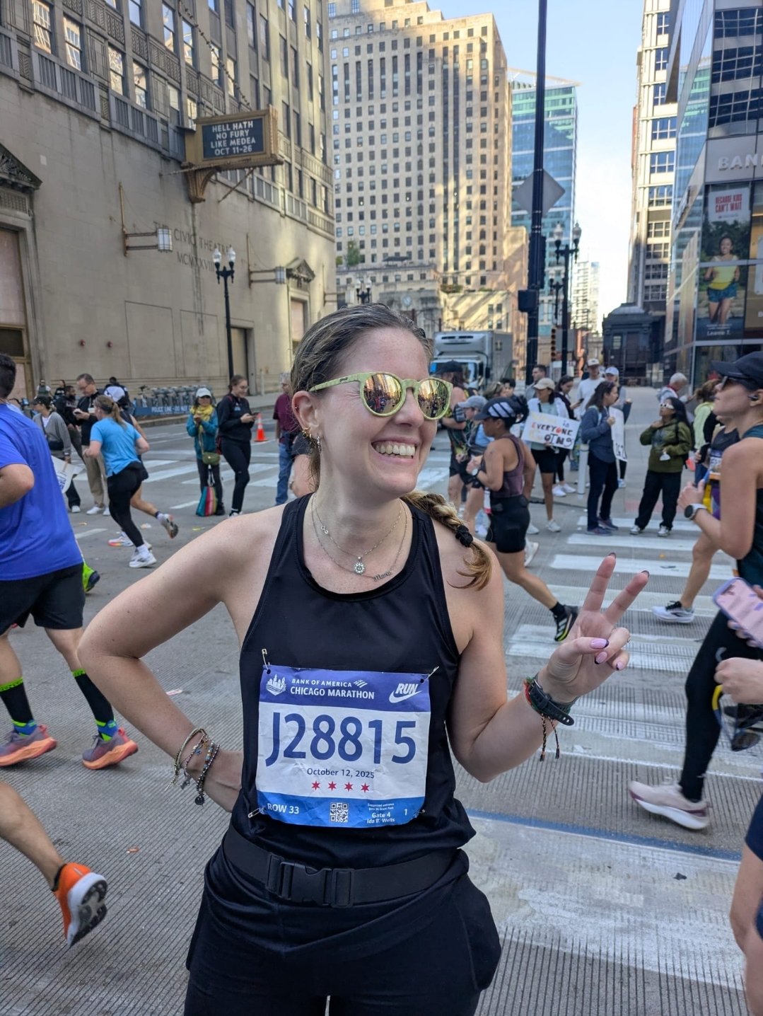 A woman smiling and making a peace sign during a city marathon, with runners and skyscrapers in the background.