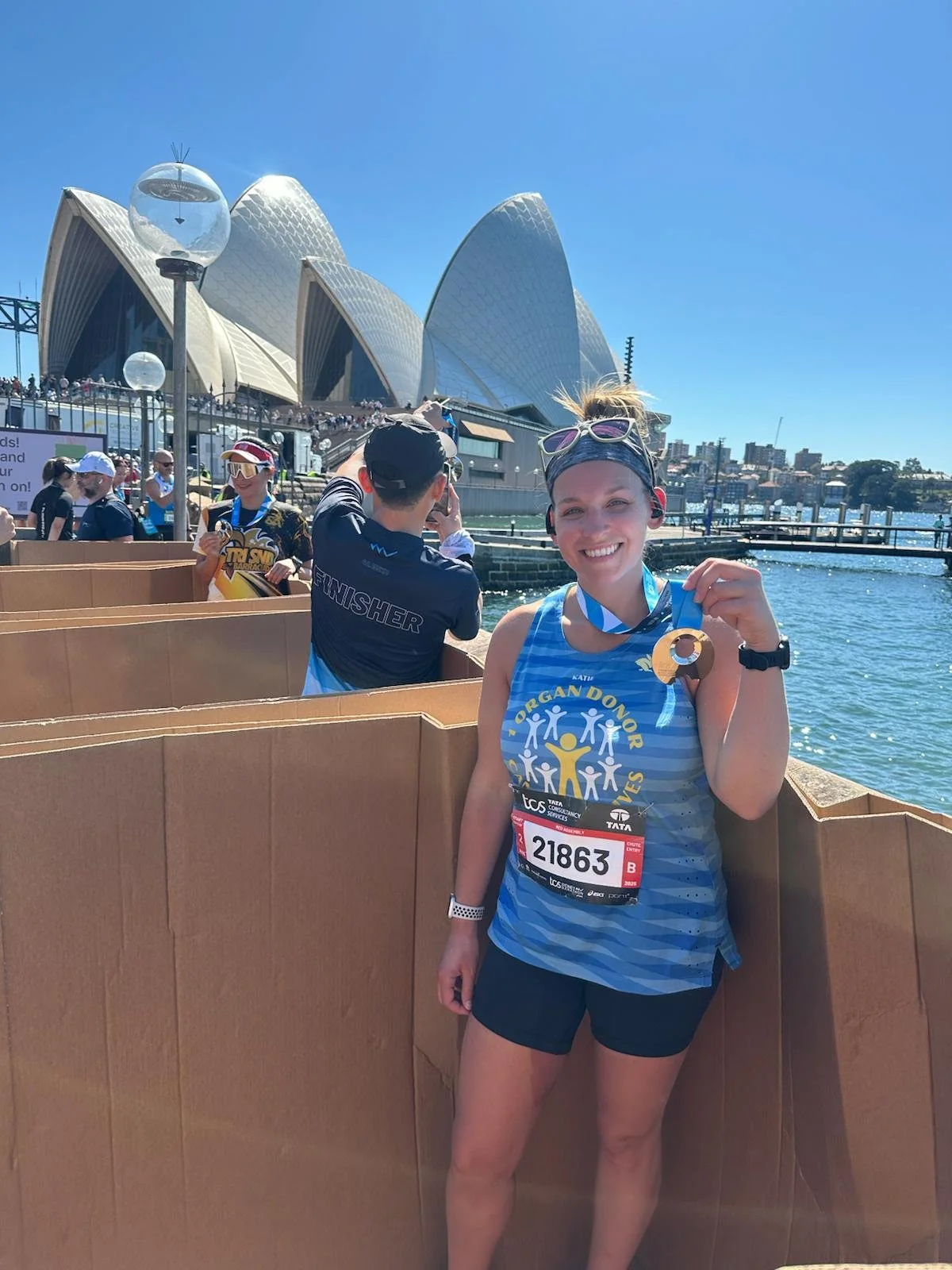 A woman smiling and holding a medal at a finish line in front of the Sydney Opera House after a race.