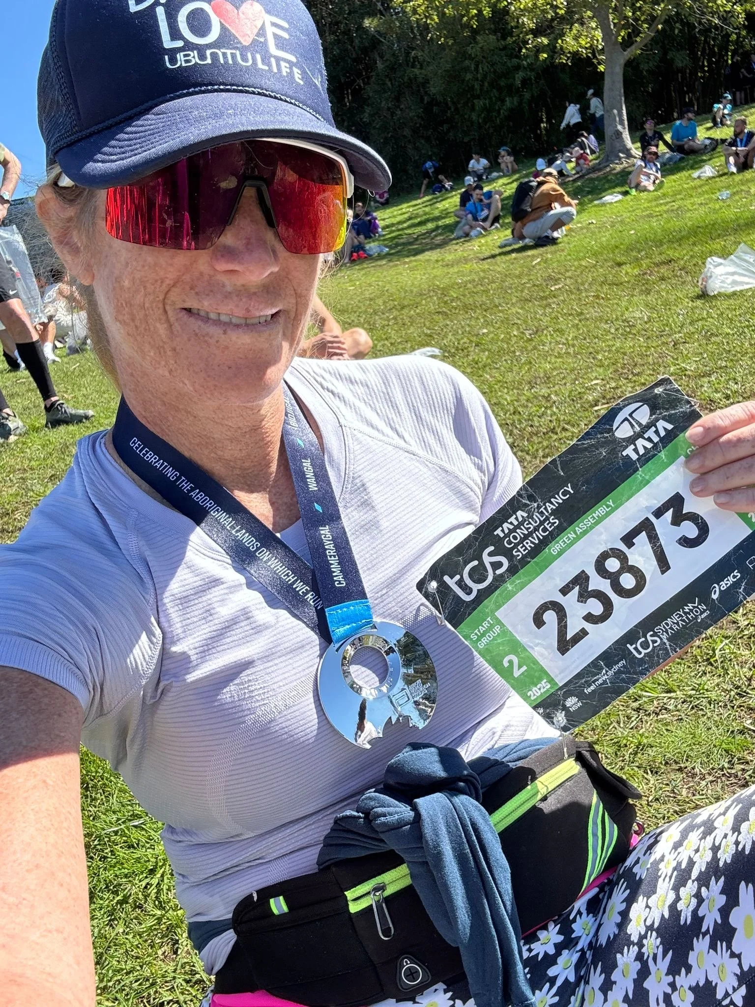 Woman taking a selfie after completing a race, wearing sunglasses, a blue cap, a medal around her neck, and holding a race bib with the number 238873 at a park with other people in the background.