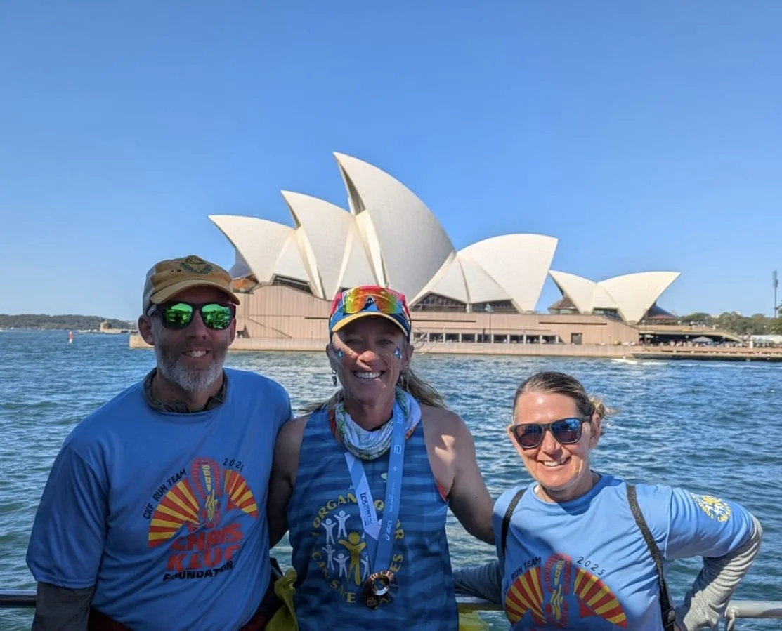 Three people in athletic gear standing in front of the Sydney Opera House on the water, smiling at the camera.