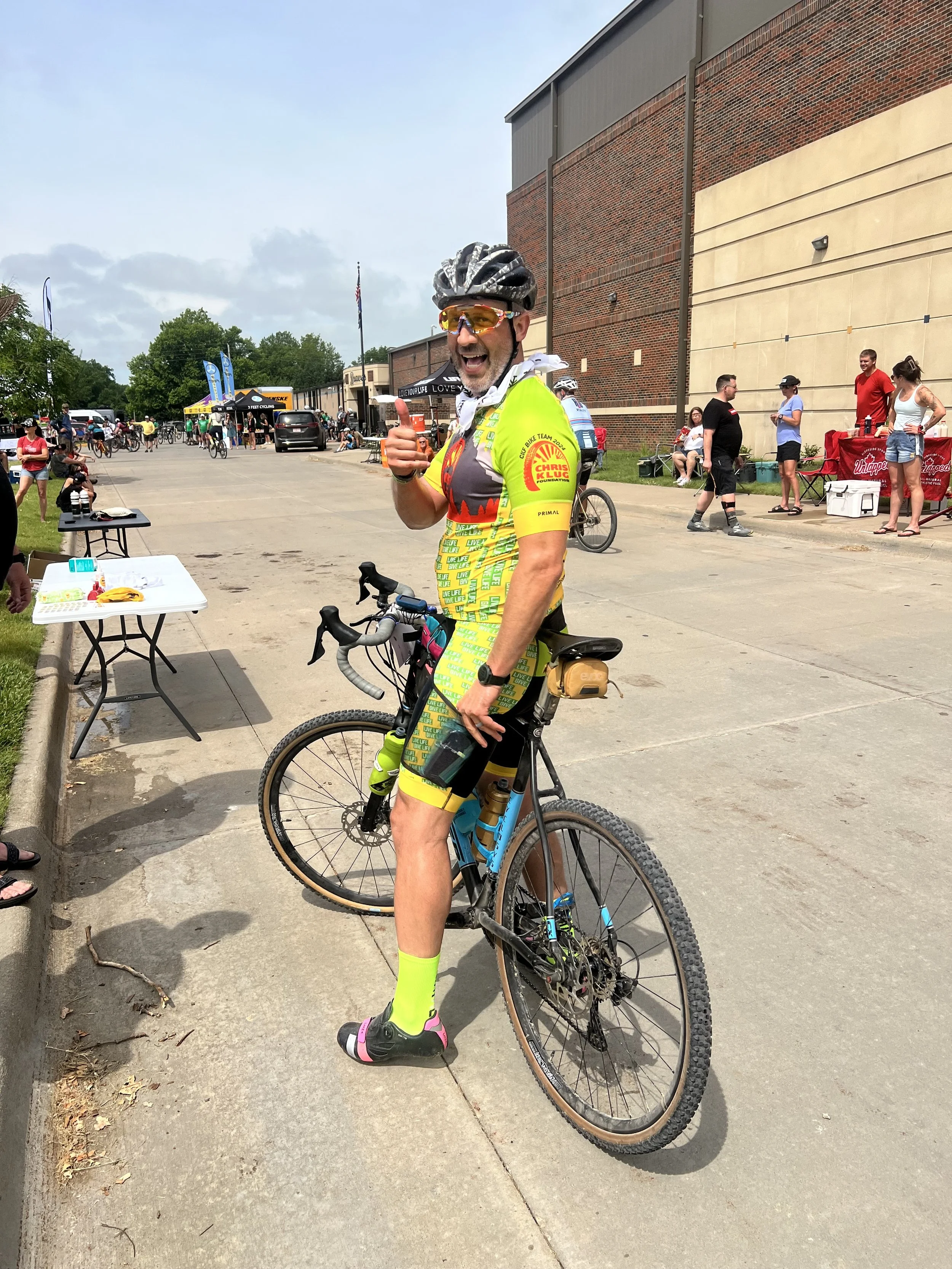 A man wearing a cycling helmet, sunglasses, and a colorful cycling jersey gives a thumbs-up while standing next to his bike at an outdoor event. Other people are in the background, some sitting at tables and some walking or riding bikes.