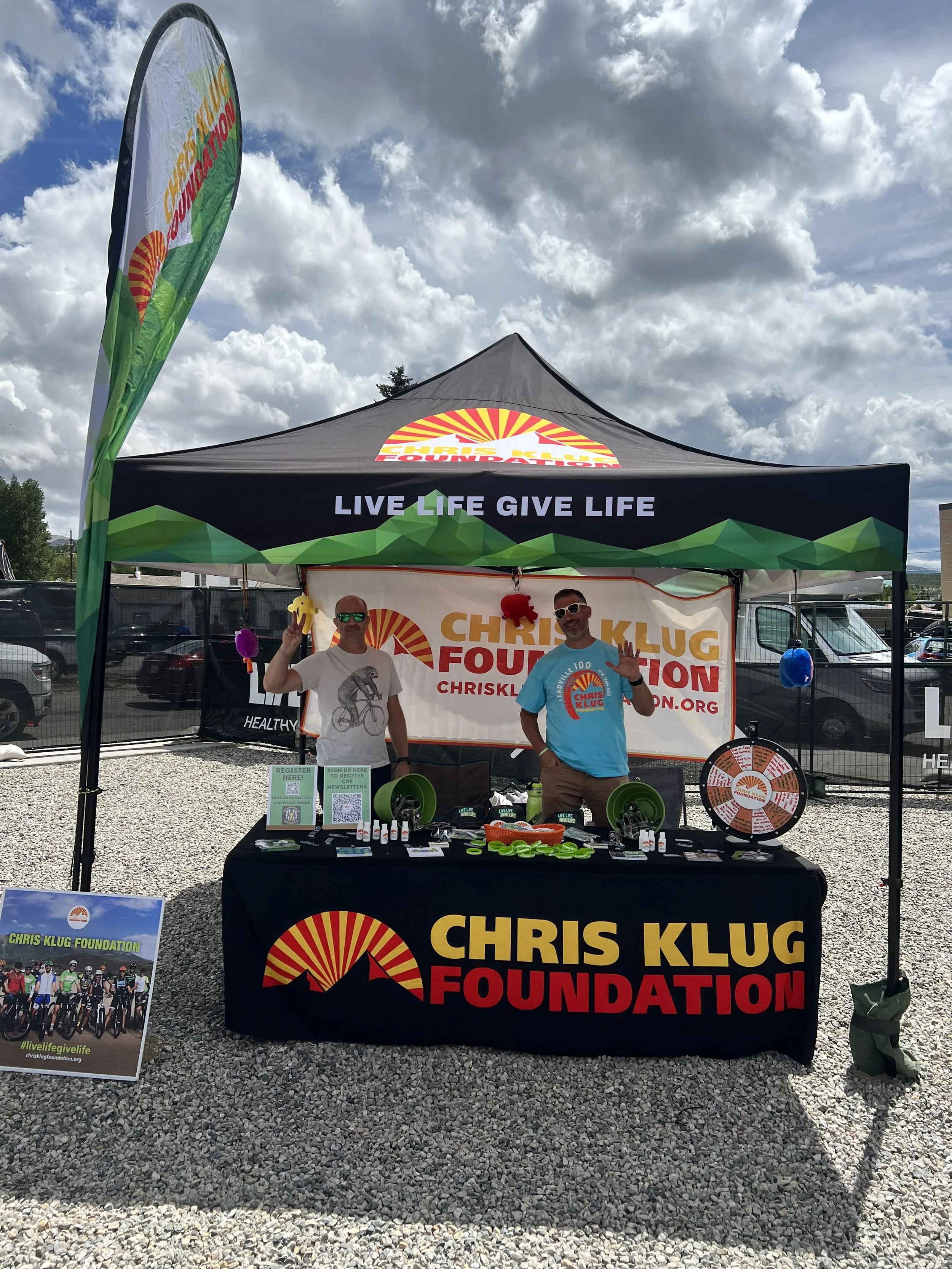 Two men standing behind a table at an outdoor event booth for the Chris Klug Foundation. The booth is set up under a black canopy tent with the foundation's logo and slogan, 'LIVE LIFE GIVE LIFE.' One man is wearing a white t-shirt and sunglasses, holding a yellow toy figure, and the other is wearing a light blue t-shirt and white sunglasses, waving. The table has promotional items, and there is a spinning prize wheel and various colorful items on display. A stand nearby features a poster with a group of cyclists and the foundation's hashtag, #livelifegivelife.