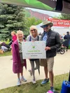 Three people smiling at a donation event outdoors, holding a sign that says, 'I SAID YES TO organ, eye, and tissue donation,' with trees, tents, and mountains in the background.