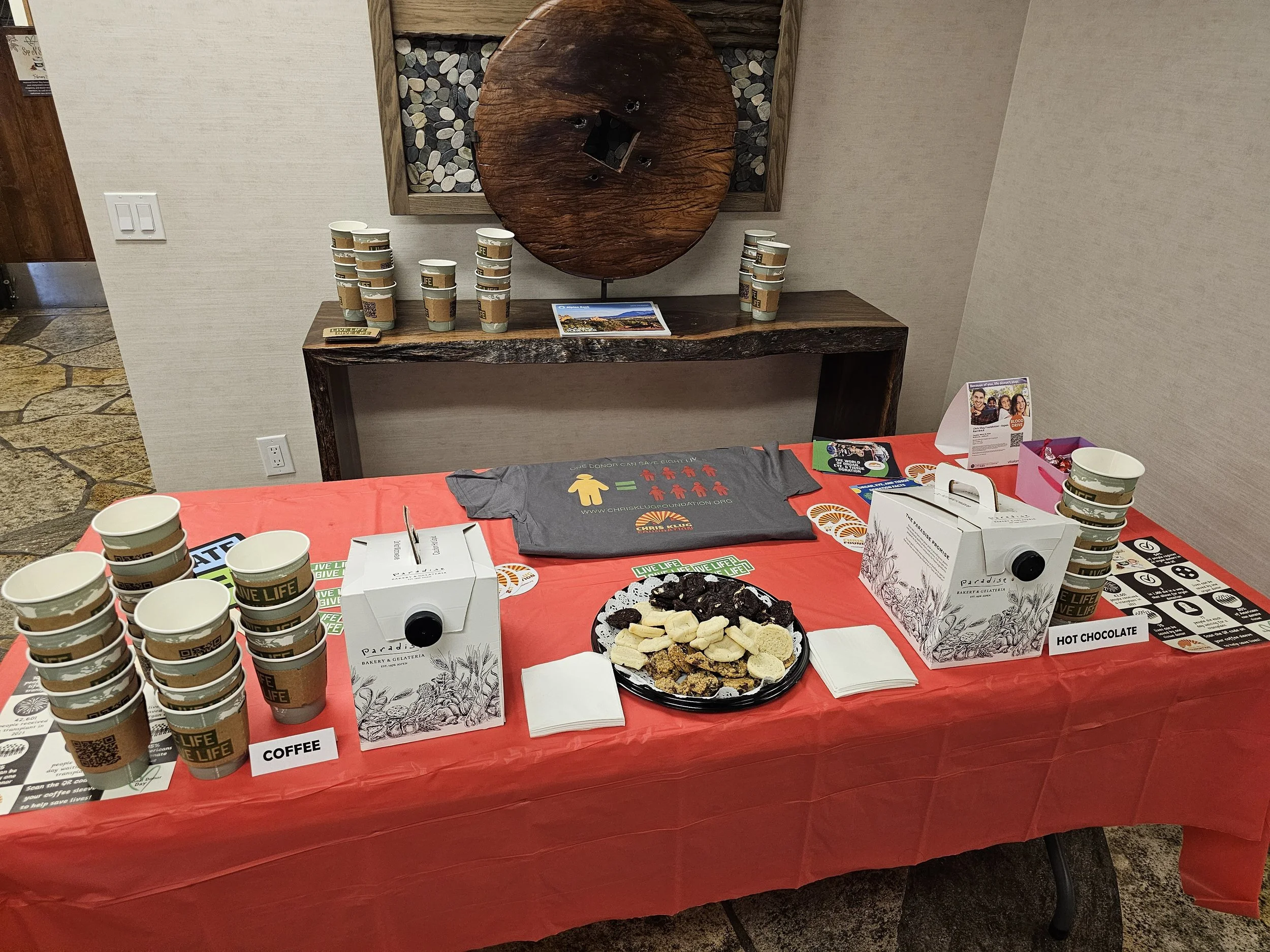Table with coffee cups, hot chocolate, snacks, and promotional material at an event, with a wooden wall and decorative artwork in the background.