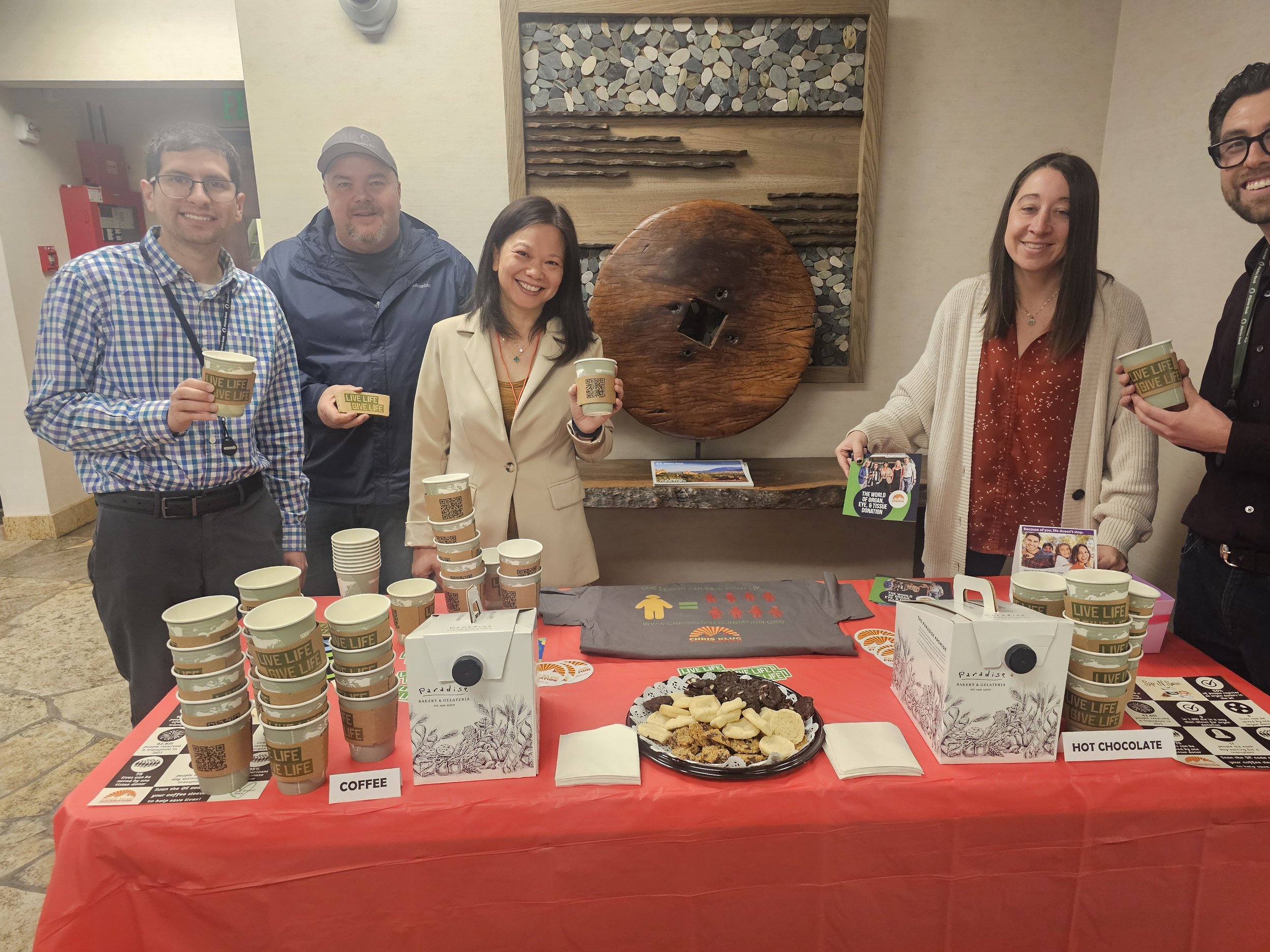Six people standing behind a table with coffee cups and snacks at an indoor event. Four are holding cups, smiling at the camera. The table has a red tablecloth, cups labeled "Live Life, Give Life," and snacks in the center. Behind them is a decorativ