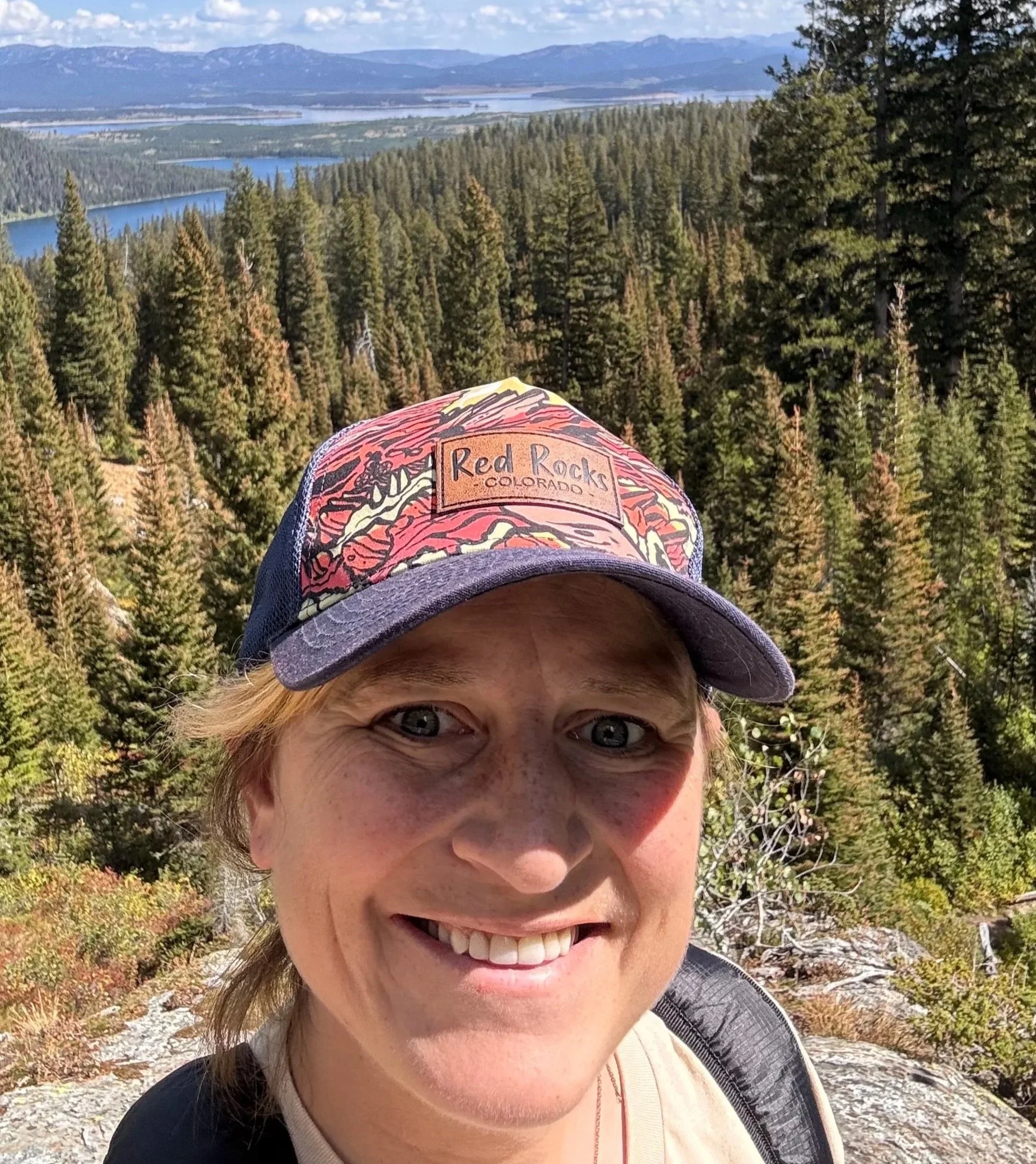 A woman taking a selfie in a mountainous forest landscape with trees, lakes, and mountains in the background on a clear day.