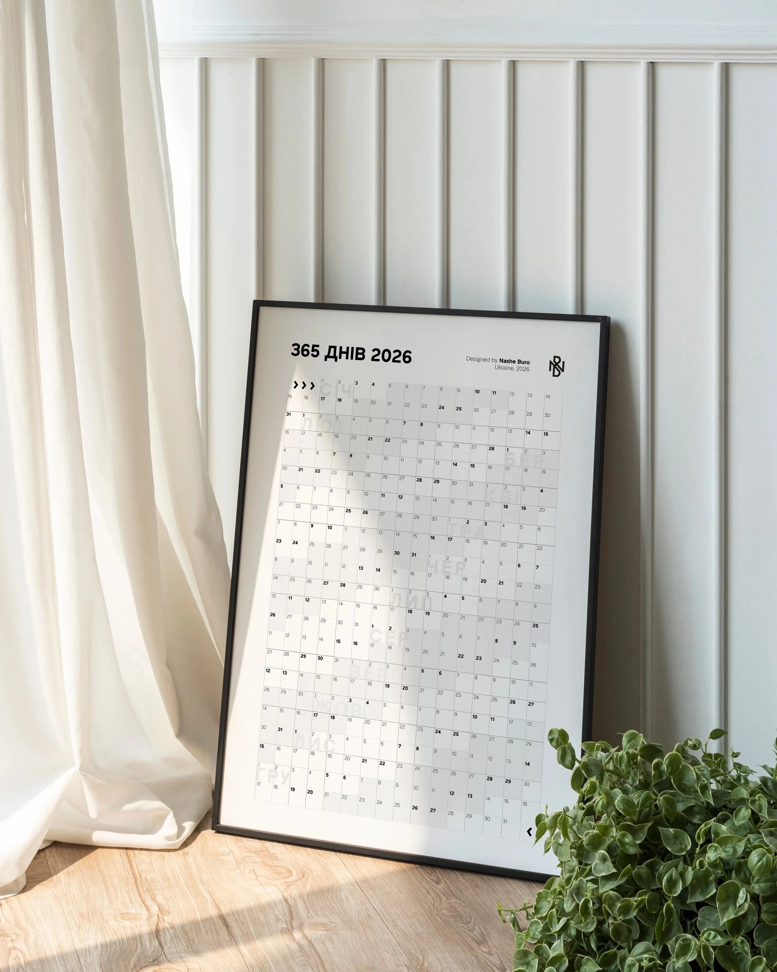 Framed 2026 calendar titled '365 ДНІВ 2026' leaning against a white wall on a wooden floor, with a potted green plant to the right and a white curtain on the left.