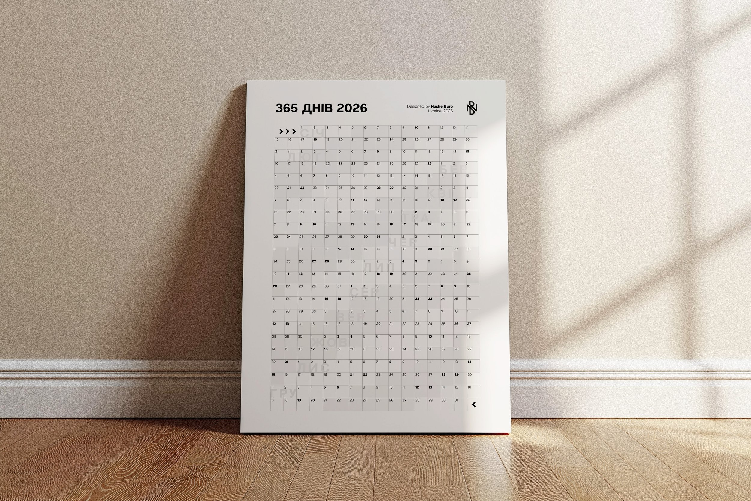 A wall calendar for the year 2026 leaning against a beige wall on a wooden floor. The calendar displays the months and dates in a grid format with text in Ukrainian.