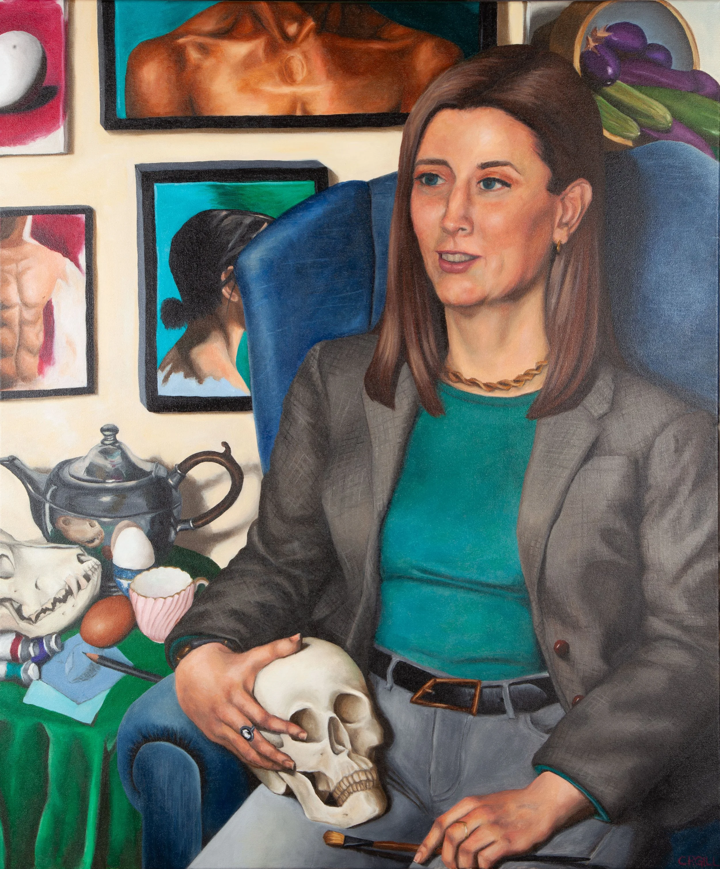A woman with long brown hair in a teal shirt and grey blazer, holding a skull and a paint brush, sits on  in front of a collection of paintings. Tea, eggs, paint, drawings, and an animal skull is on the table beside her.