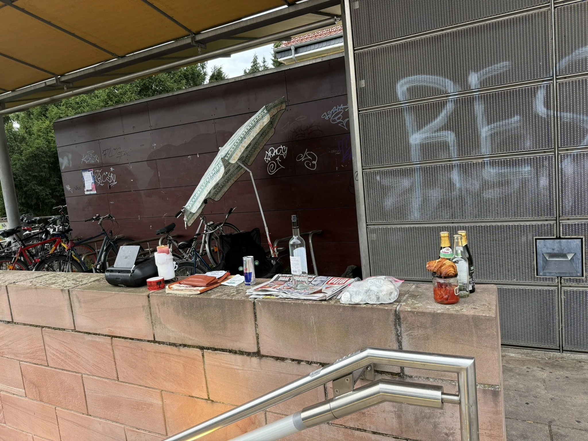 Auf der Balustrade am Treppenabgang stehen Getränke, ein Croissants, eine Tageszeitung und ein Sonnenschirm ordentlich aufgereiht