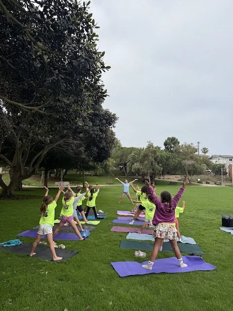 Children participating in an outdoor yoga class on a grassy park area, led by an instructor, with trees and school in the background.