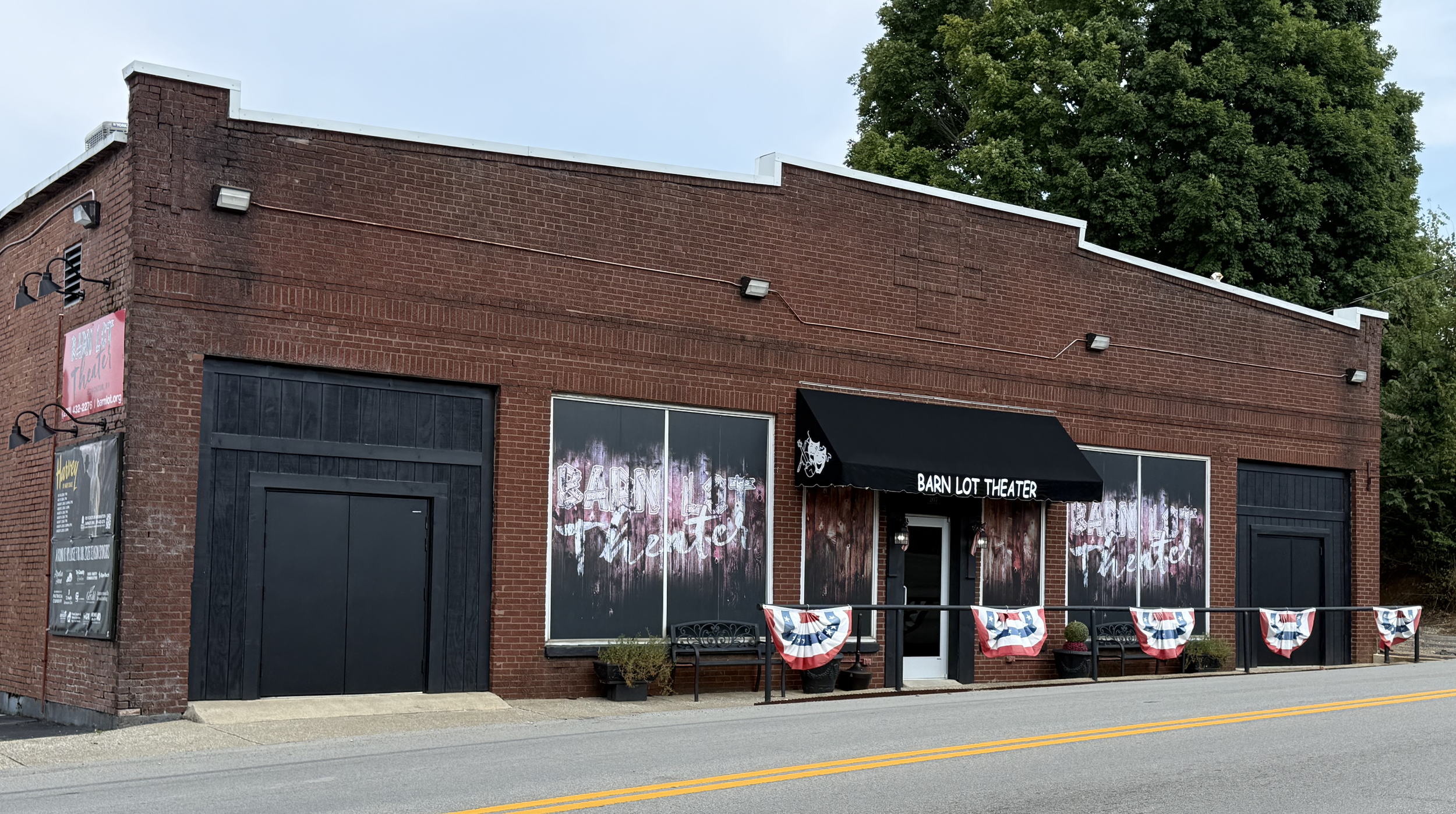 Brown brick building with black double doors on each side of the building. A central glass door under a black awning that reads Barn Lot Theater. Red and black signage are hanging on the side of the building.
