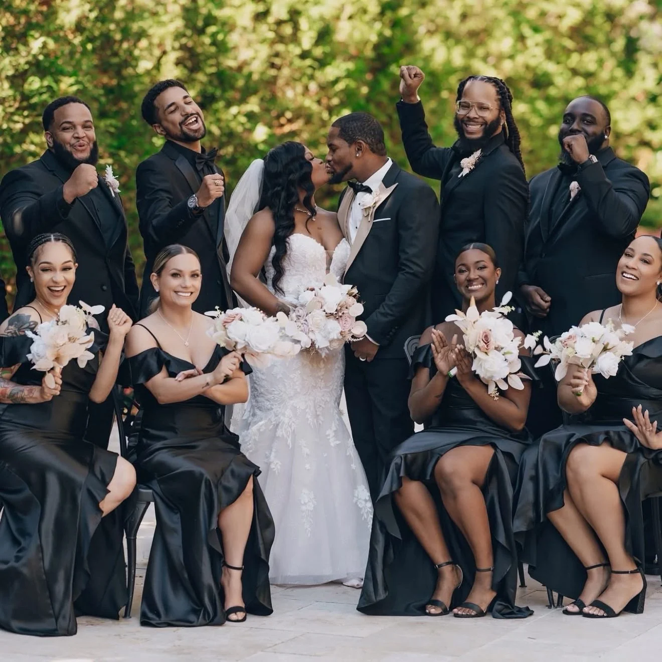 Wedding party group photo — bride and groom kissing, bridesmaids in black dresses